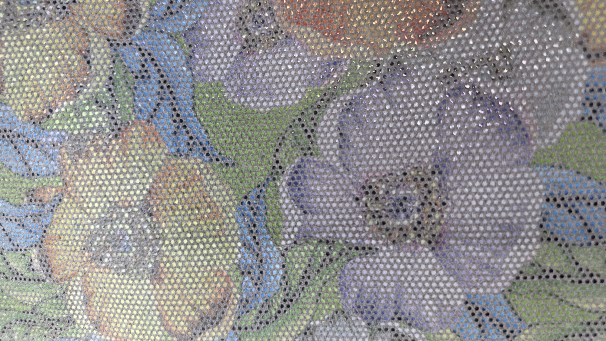 The image is a close-up, slightly blurry shot of a patterned fabric. The pattern appears to be floral, with large, abstract shapes in shades of purple, yellow, orange, blue, and green. The fabric itself has a textured appearance due to a visible grid of small, circular perforations. Some of the perforations reflect light, giving the fabric a subtle shimmer or glittery effect. The background is indistinct due to the close-up view and the texture of the fabric. There are no people or identifiable objects within the frame, and the context suggests it is a close-up of material. There is no information that can be used to determine a location in Birdik, Kyrgyzstan.