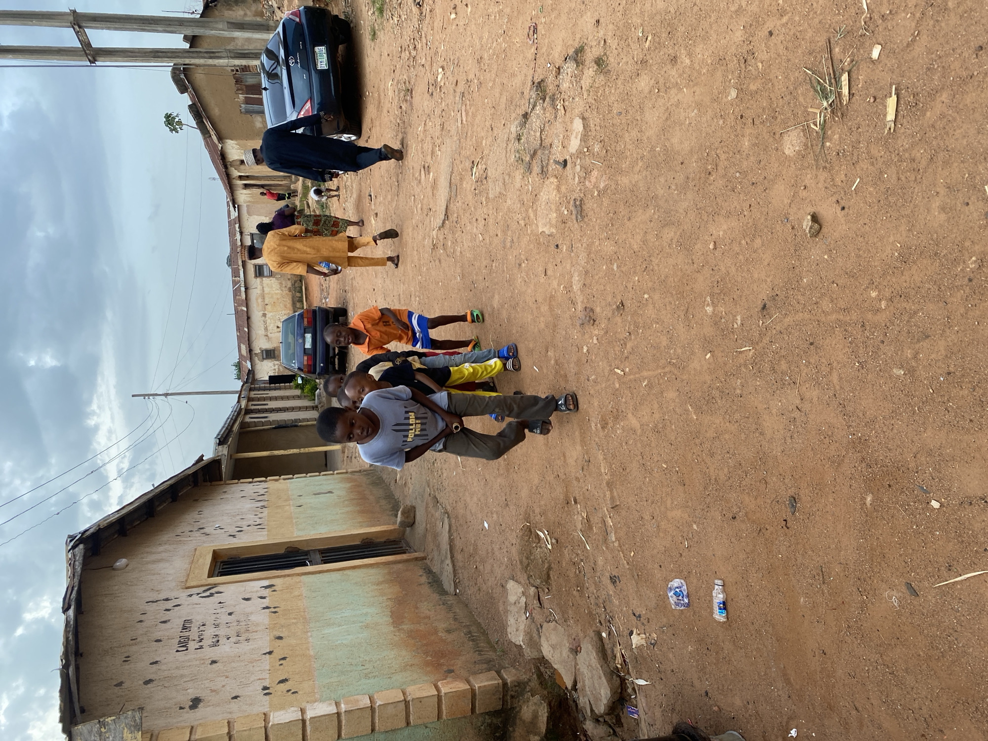 In a dusty, unpaved street in Bukuru, Nigeria, a group of children walk in a line. The child in the foreground, wearing a light blue t-shirt with the text "FOLLOW ME!", is looking towards the camera. Behind him, three other children follow, two of whom are looking ahead and one is looking to the side. One of the children is wearing a yellow t-shirt and blue shorts.
The street is lined with modest, single-story buildings constructed from materials like concrete blocks and corrugated metal. Some of the buildings appear weathered, with peeling paint and visible repairs. One building on the left has a sign with text that is partially visible, reading "CAVEAT EMPTOR".
Two cars are parked in the background, a dark blue sedan and a lighter colored sedan. Several adults are also present in the scene, including a man in a yellow robe and another in a dark robe, walking away from the camera. The sky above is overcast with grey clouds. Scattered on the ground are various pieces of debris, including plastic bottles. The ground itself is a reddish-brown dirt, indicative of the Nigerian soil.