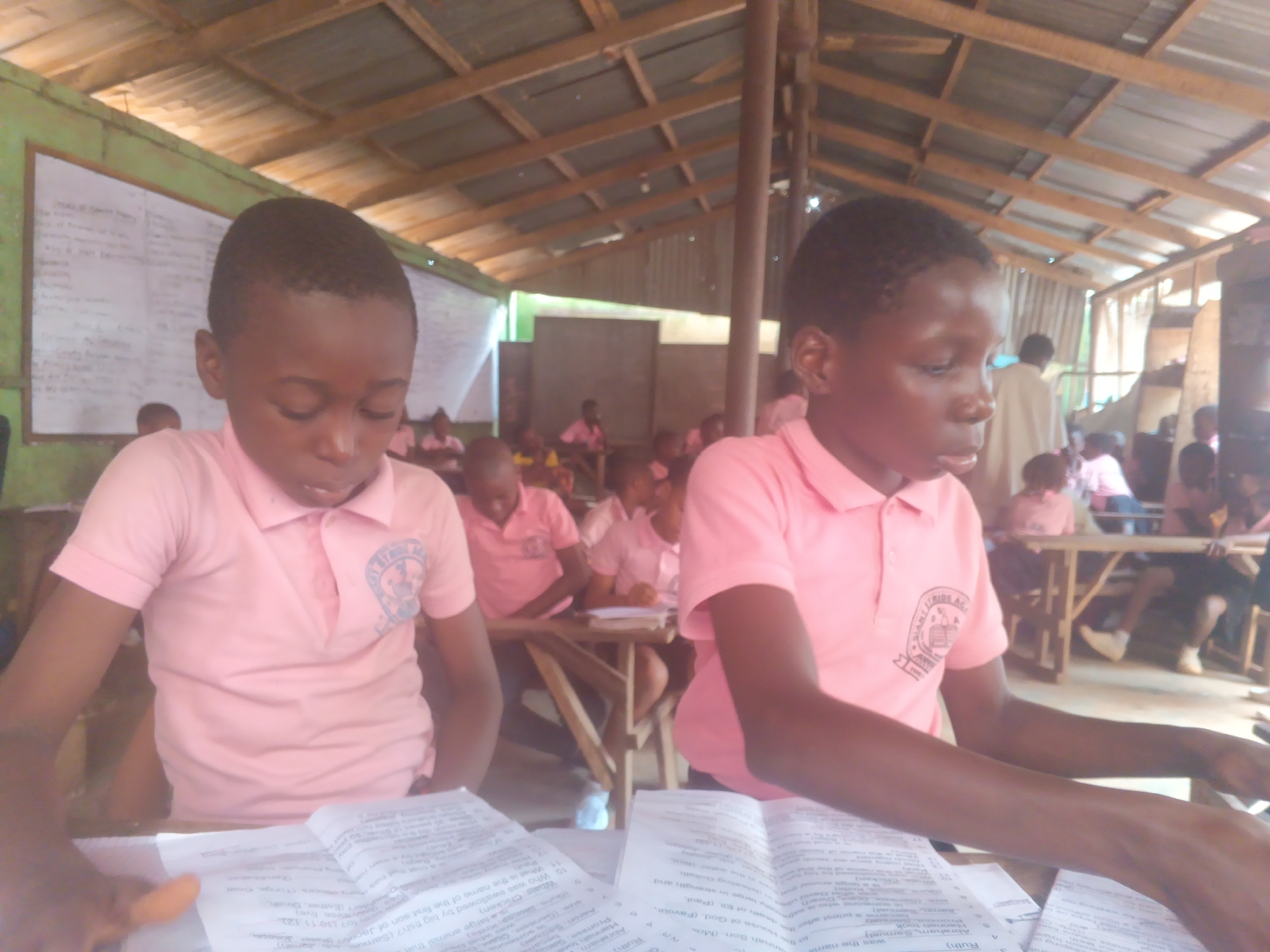 The image depicts a classroom scene in Onitsha, Nigeria, characterized by a rustic, open-air structure with a corrugated metal roof supported by wooden beams. In the foreground, two young students, a boy on the left and a girl on the right, are seated at a wooden table. Both are wearing pink polo shirts with the emblem "Saint Jude Academy" visible on the boy's shirt. They are focused on papers spread out before them, appearing to be engaged in schoolwork or an examination. The boy on the left is leaning over his papers, while the girl on the right has her hands resting on her papers, suggesting she is reading or writing.
The background reveals more students in similar pink attire, seated at other tables and desks within the classroom. The environment appears to be a humble educational setting, possibly a primary or secondary school, with visible chalkboards and posters on the walls, though details are indistinct. The overall impression is of a busy, active learning environment in a developing urban area.