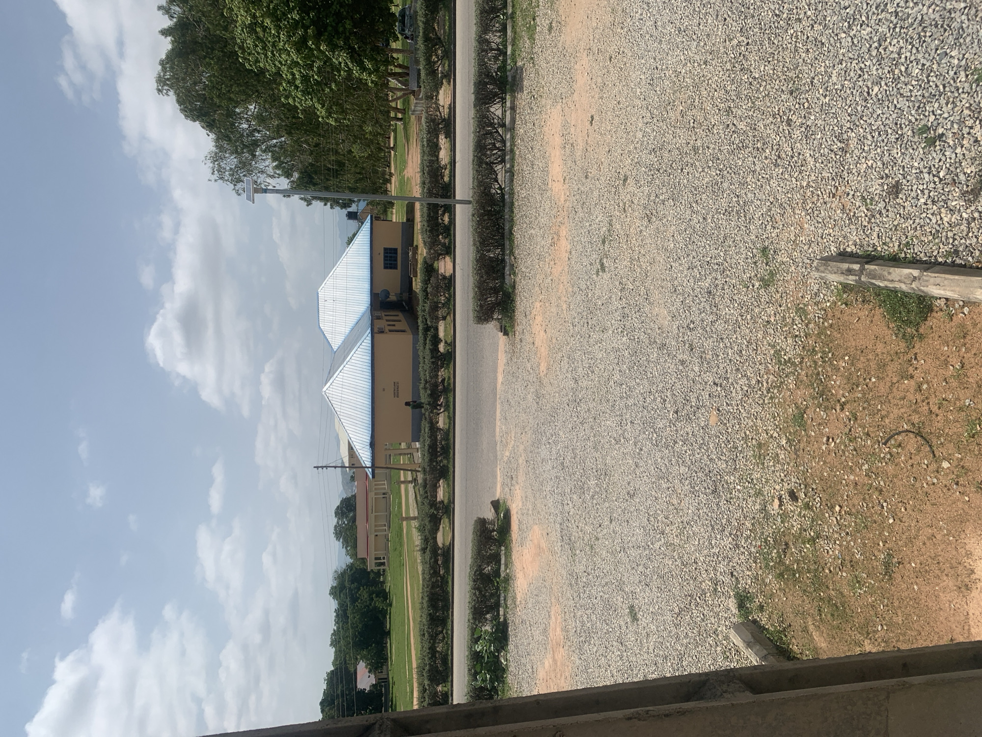The image captures an outdoor scene on a bright, cloudy day in Jalingo, Nigeria. In the mid-ground, two distinct buildings with light blue, corrugated metal roofs are visible. The buildings are yellow with dark trim around the windows. One person, dressed in dark clothing, is standing near the entrance of the right-hand building. A paved road runs in front of the buildings, bordered by neatly trimmed hedges. A tall, thin pole with a solar panel on top stands behind the hedges, to the right of the buildings. Beyond the road and buildings, a green field stretches out, with several trees and other, less distinct structures in the distance. The foreground of the image is dominated by a gravel-covered area, with patches of dry, reddish-brown earth and sparse green grass. To the left, a concrete pillar or beam is visible, suggesting the edge of a structure or pathway. The overall environment appears to be an institutional or campus setting, possibly a school or administrative complex, given the layout and presence of multiple buildings.
