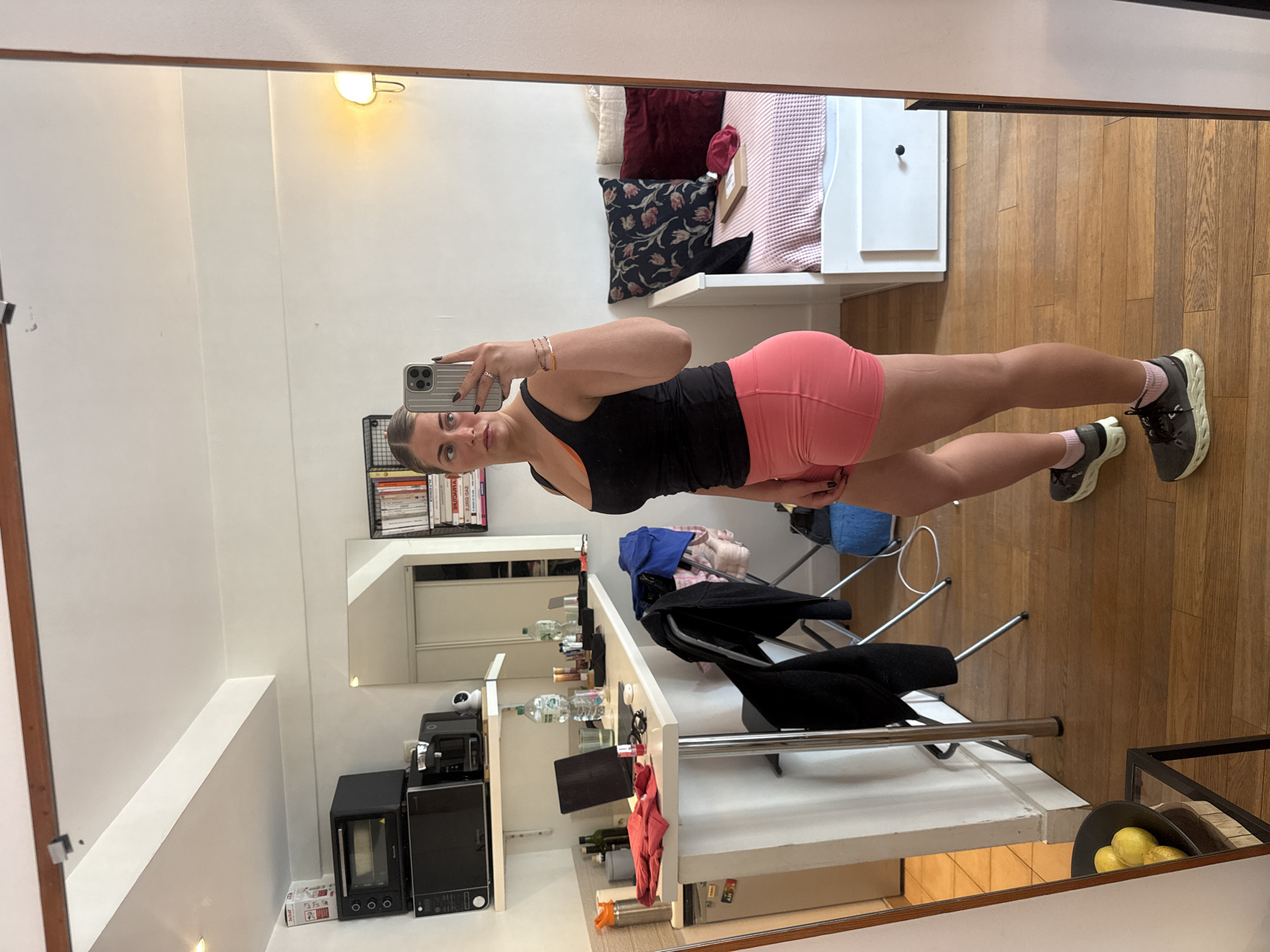 A young adult woman takes a mirror selfie indoors. She wears a black tank top and pink shorts, as well as grey sneakers with pink socks. The room includes a white shelf, a microwave oven, a small couch with pillows, a wooden floor, and a table with various items on it. Based on the architectural style and decor, the location could be a compact living space in an urban setting. Without further information, it is impossible to confirm it is in Paris, France.