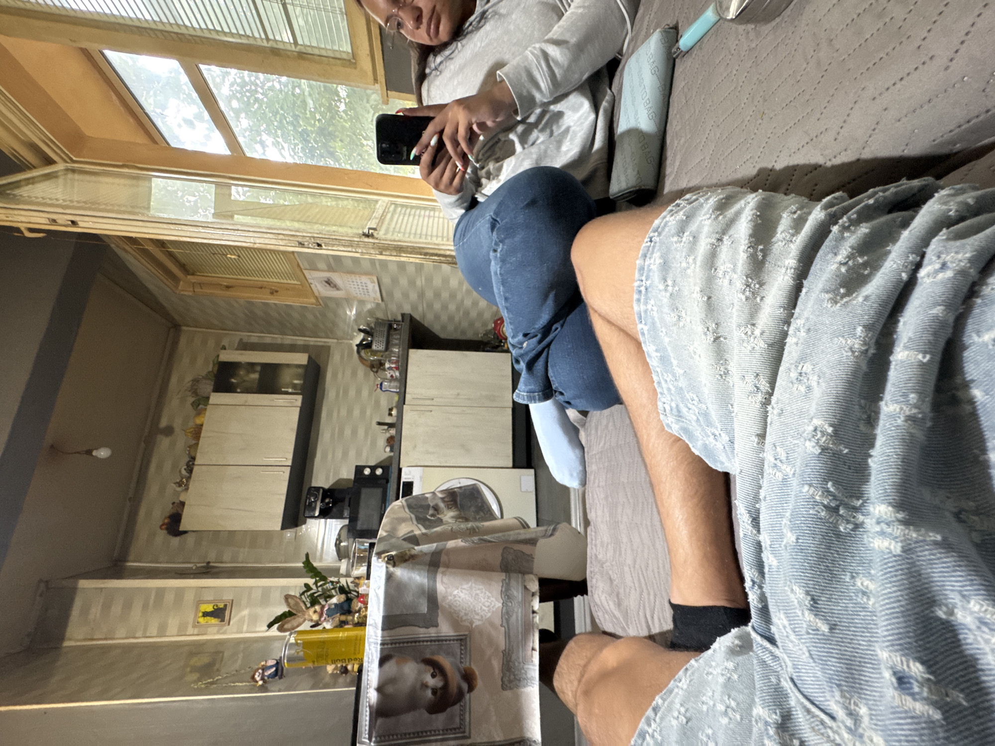 In a residential apartment in Sofia, Bulgaria, a woman reclines on a bed, holding a smartphone and looking at its screen. She is wearing a gray hooded sweatshirt and blue jeans. Beside her, a man's legs are visible, wearing denim shorts and black socks. The background reveals a small kitchen area with cabinets, a microwave, and a washing machine. A table covered with a patterned cloth is also visible, adorned with decorative items. The room has a large window with blinds, offering a view of trees outside. The overall setting suggests a casual, domestic environment within an apartment.