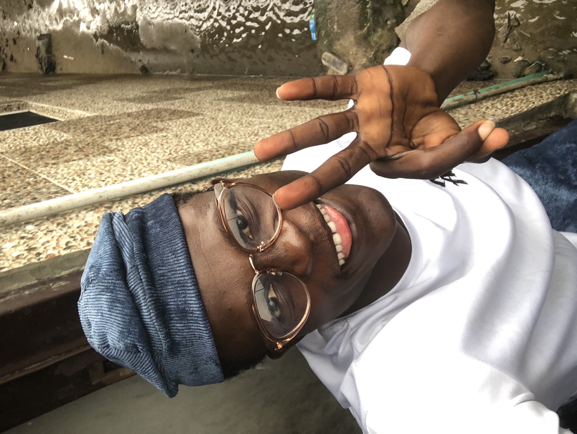 In Ado, Nigeria, a young Black man wearing a blue corduroy cap and glasses smiles broadly at the camera, sticking out his tongue. He is dressed in a white t-shirt and blue jeans. He holds up two fingers in a peace sign. The background is a textured wall with some greenish pipe and a dirty, wet ground surface with some water flowing. The lighting is bright, suggesting it's daytime.