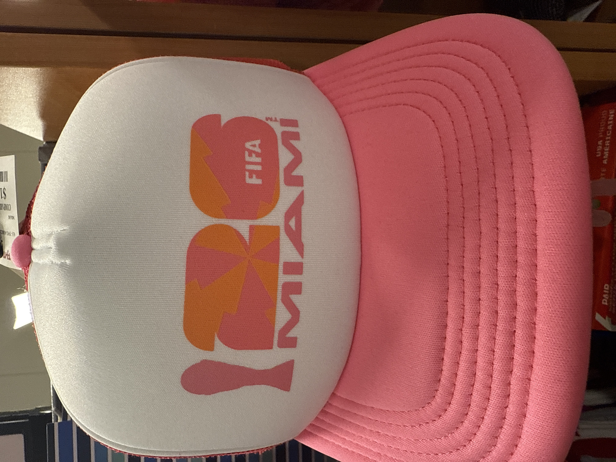The image shows a pink and white trucker hat. The front of the hat is white with the words "2026 FIFA MIAMI" printed on it in pink and orange. The brim of the hat is pink. The hat is resting on a shelf. The location is Dania Beach, United States of America.