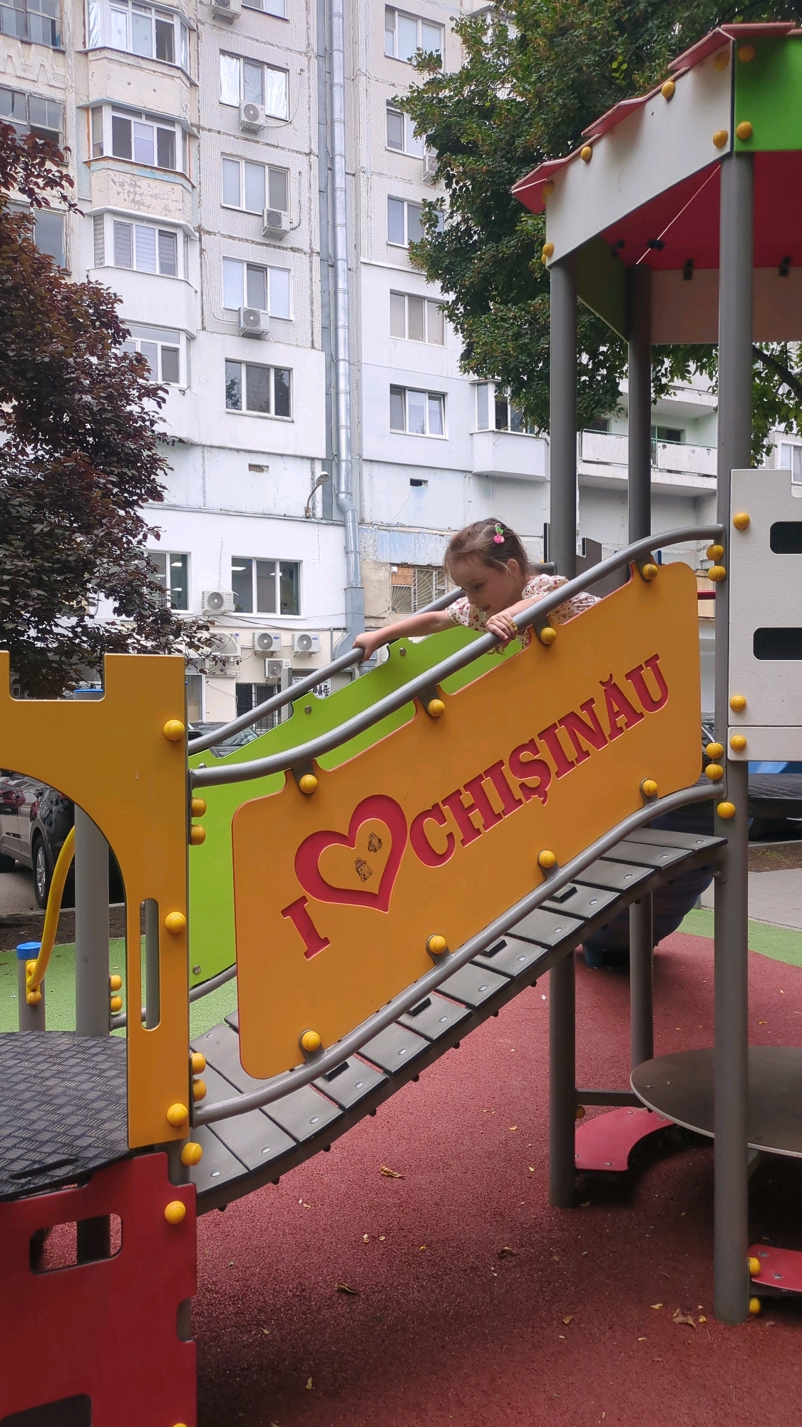 A young girl with a pink bow in her hair is on a playground, leaning on a railing. She is wearing a floral print shirt. The playground features a bright yellow slide with the words "I ❤️ CHIȘINĂU" in red letters. Behind the playground, there is a light-colored building with multiple windows and air conditioning units. Trees with dark red and green leaves are also visible. The ground is covered in a red, rubberized surface typical of playgrounds. The scene appears to be an outdoor public space in Chișinău, Moldova.
