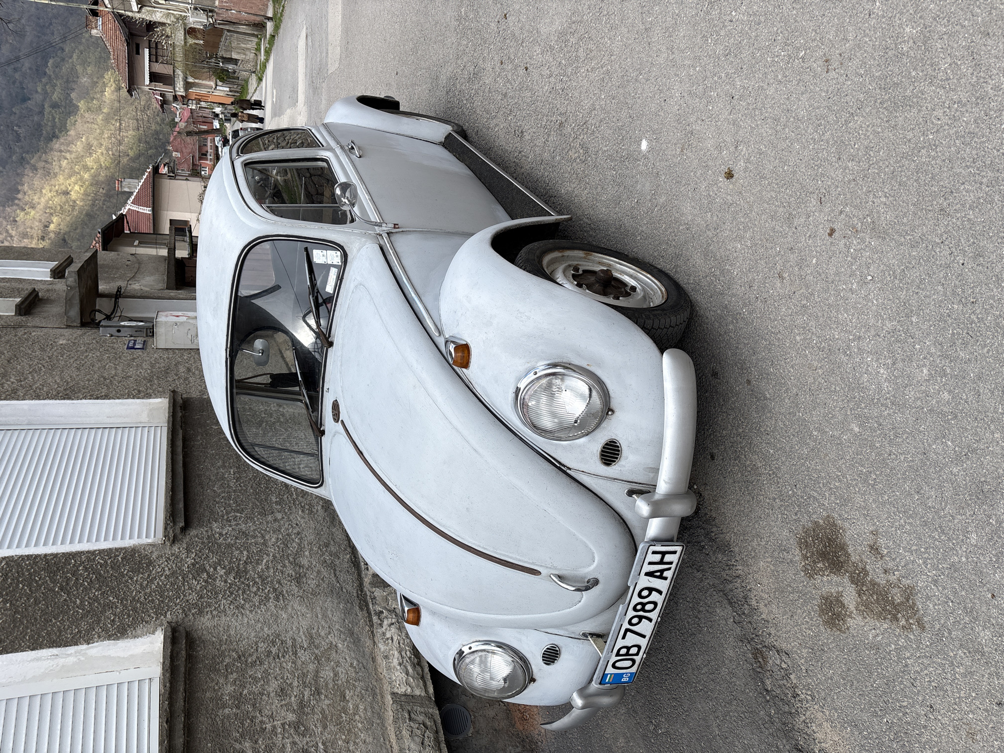 The image features a white Volkswagen Beetle parked on a street in what appears to be the city of Teteven, Bulgaria. The car is situated close to a building with white, slatted shutters and a textured gray exterior wall. The background includes a view down the street, showcasing other buildings and possibly mountainous terrain in the distance. The car's license plate reads "OB 7989 AH". The road is paved, and there are some darker marks on its surface.