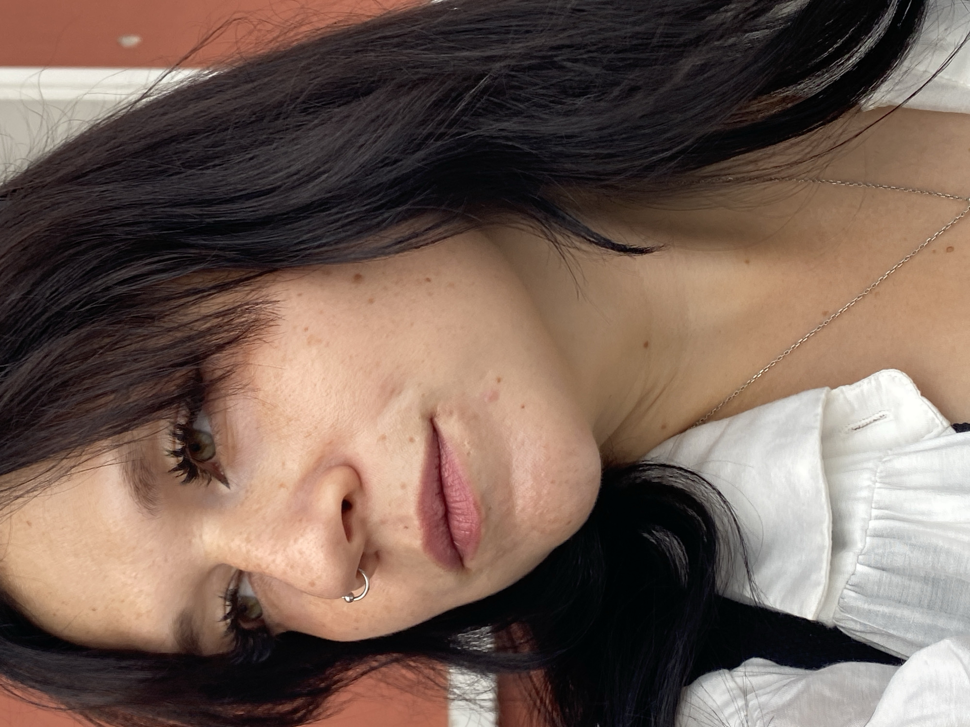 A close-up, profile shot shows a young woman with dark hair and fair skin with freckles. She is wearing a silver nose ring and a white collared shirt with ruffles. A silver necklace is visible around her neck. The background is blurred, but appears to be a red wall and white structure. No specific location in Saint Petersburg is discernible from the image.
