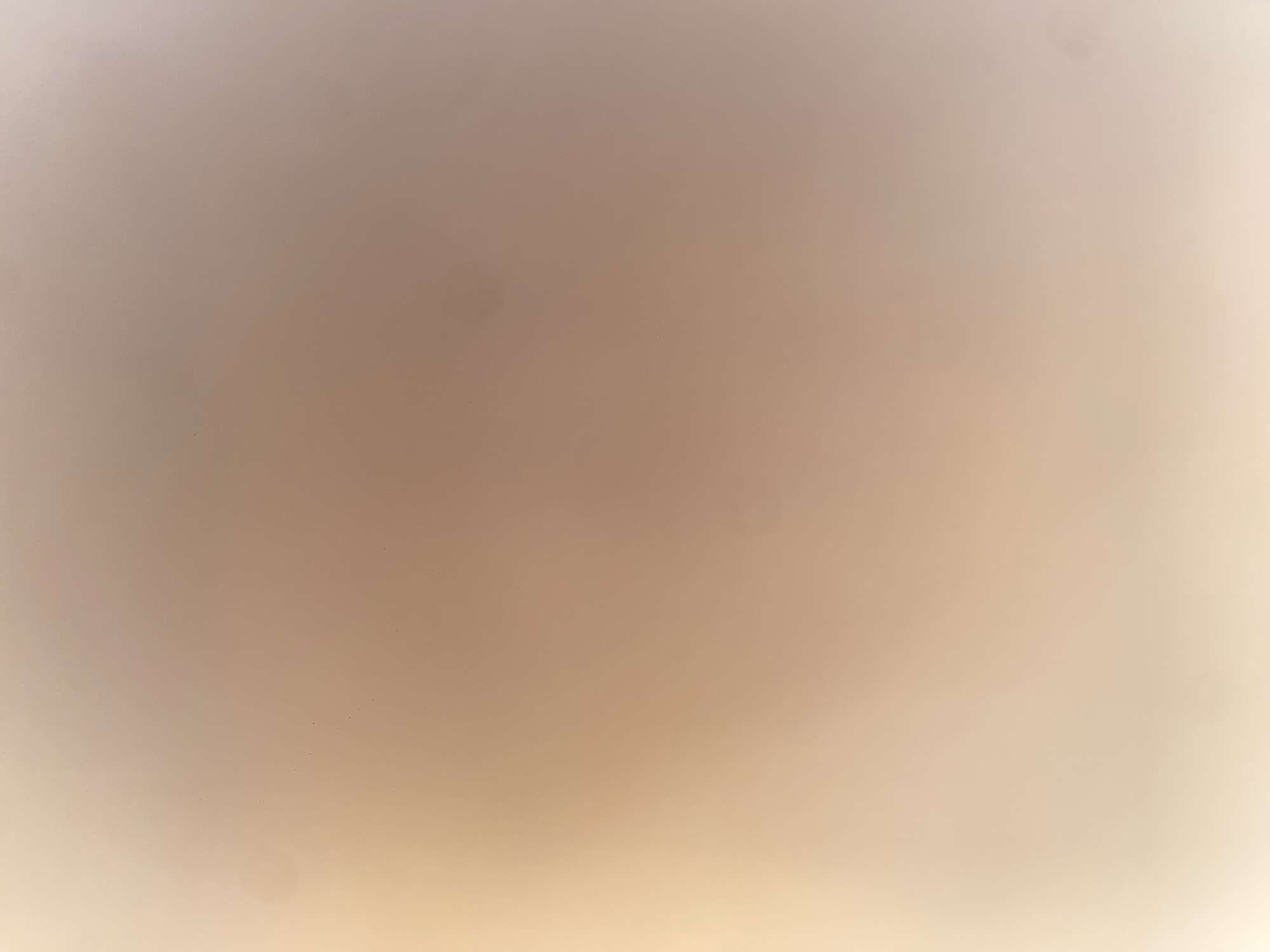This image is a close-up, out-of-focus shot of a wall in Ablanitsa, Bulgaria. The wall has a gradient of beige and peach tones, with some areas appearing lighter due to light sources. There are a few small dark specks visible on the surface, which could be dust or minor imperfections. The overall impression is of a plain, indoor wall.