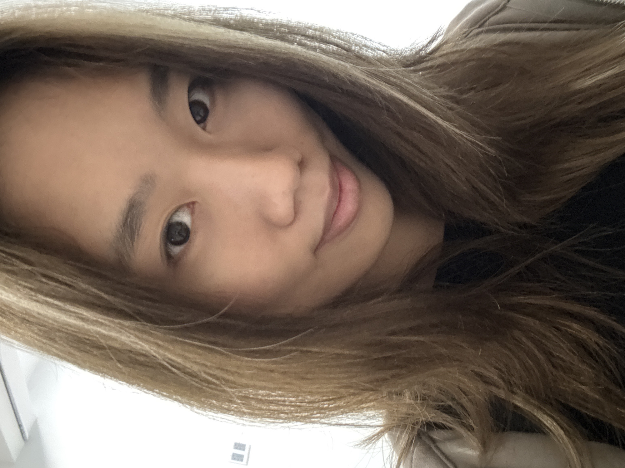 A close-up, canted head-and-shoulders shot depicts an Asian woman with shoulder-length, light brown hair. Her face is partially visible; she looks directly at the camera with a slight smile. Her expression is relaxed. She wears a dark top and what appears to be a beige jacket. The background is blurred, with a hint of white wall and a window in the lower left corner. No specific location in Nan'an District, China, is discernible from the image.