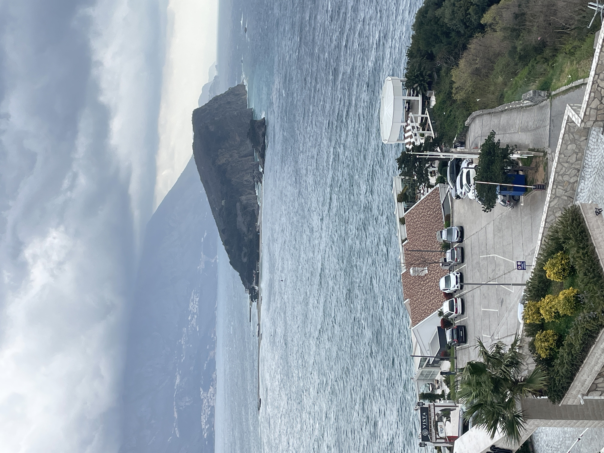 The image presents an elevated coastal view in Budva, Montenegro, under an overcast sky characterized by dense grey clouds.
In the foreground, a paved parking lot occupies the central lower portion, featuring white demarcation lines. Five vehicles are parked: a black sedan, a white SUV, a silver minivan, a grey sedan, and a white SUV. To the immediate left of the parking lot, a structure with a terracotta tiled roof and white walls is visible, displaying a vertical sign that reads "VISTA." A large palm tree stands in the lower-left, next to landscaped planters containing green and yellow shrubs. A blue parking barrier system and a winding paved road descending to the right are adjacent to the parking area. Dense green foliage covers the sloped terrain to the right of the road. Further right, a white, circular, open-air pavilion with a roof overlooks the sea.
The midground is dominated by the Adriatic Sea, displaying a rough, choppy surface with numerous whitecaps, indicating strong wind or waves. A long, narrow landmass or breakwater extends into the sea from the mid-left. A single red buoy is present in the water.
In the background, the prominent, rocky Sveti Nikola Island rises from the sea, covered with dark green vegetation. Further along the distant coastline, additional rugged cliffs and smaller land formations are visible. Distant mountains are partially obscured by cloud cover on the far left horizon.