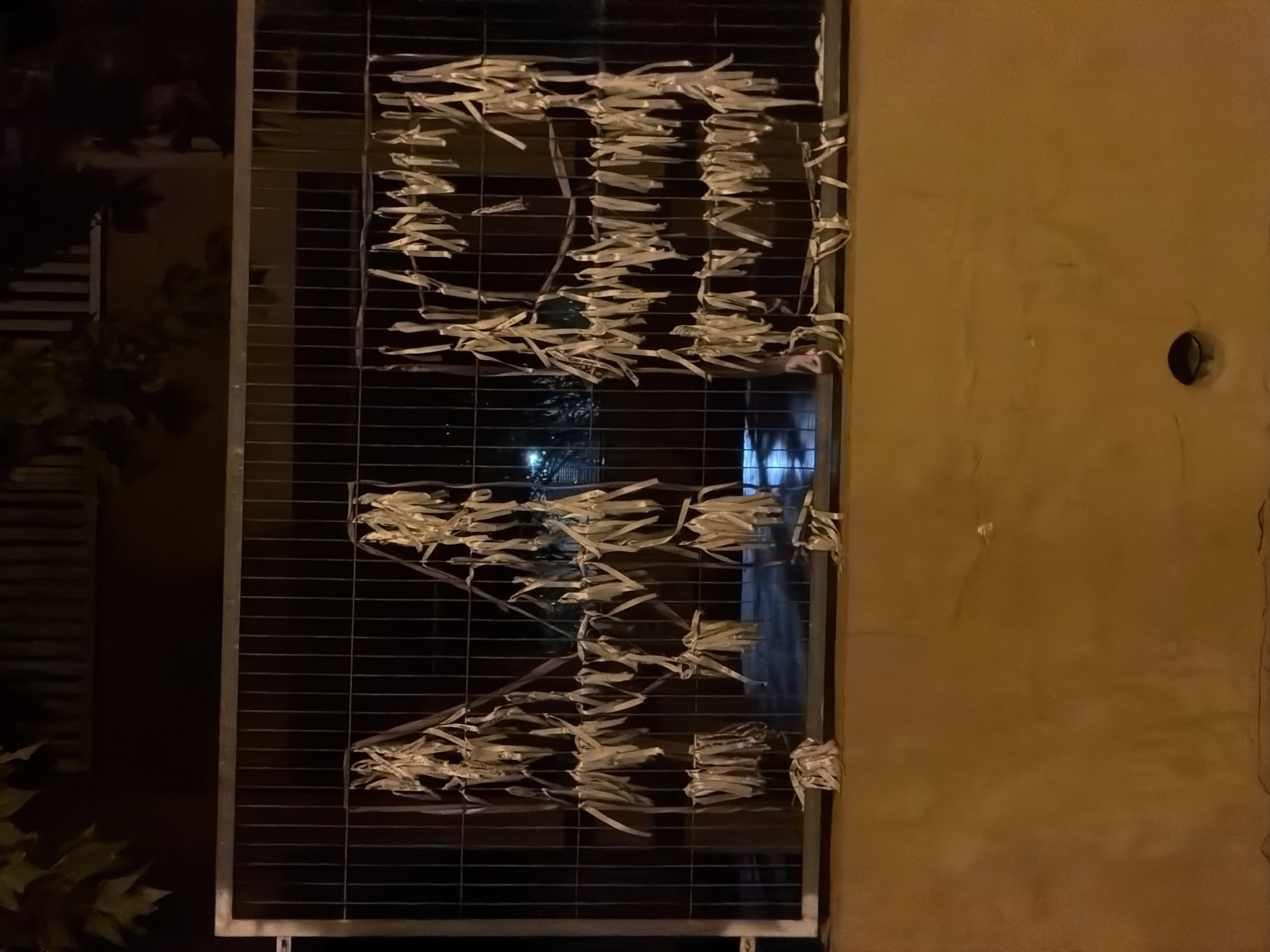 An outdoor nighttime photograph captures a section of a chain-link fence adorned with numerous hanging strips of white and light-colored material, possibly fabric or paper. These strips are arranged in clusters, creating a decorative or symbolic effect across the fence. The fence itself is silver-colored and appears to be part of a temporary barrier, as suggested by its construction.
Below the fence is a rough, textured wall, painted in a warm, earthy orange tone. The wall has a visible drainpipe opening near its base. In the background, dark foliage can be seen above the fence, and beyond that, hints of buildings and streetlights suggest an urban or suburban setting. The lighting is artificial and casts shadows, contributing to the nocturnal atmosphere.
The image is believed to have been taken in the city of Calvià, Spain.