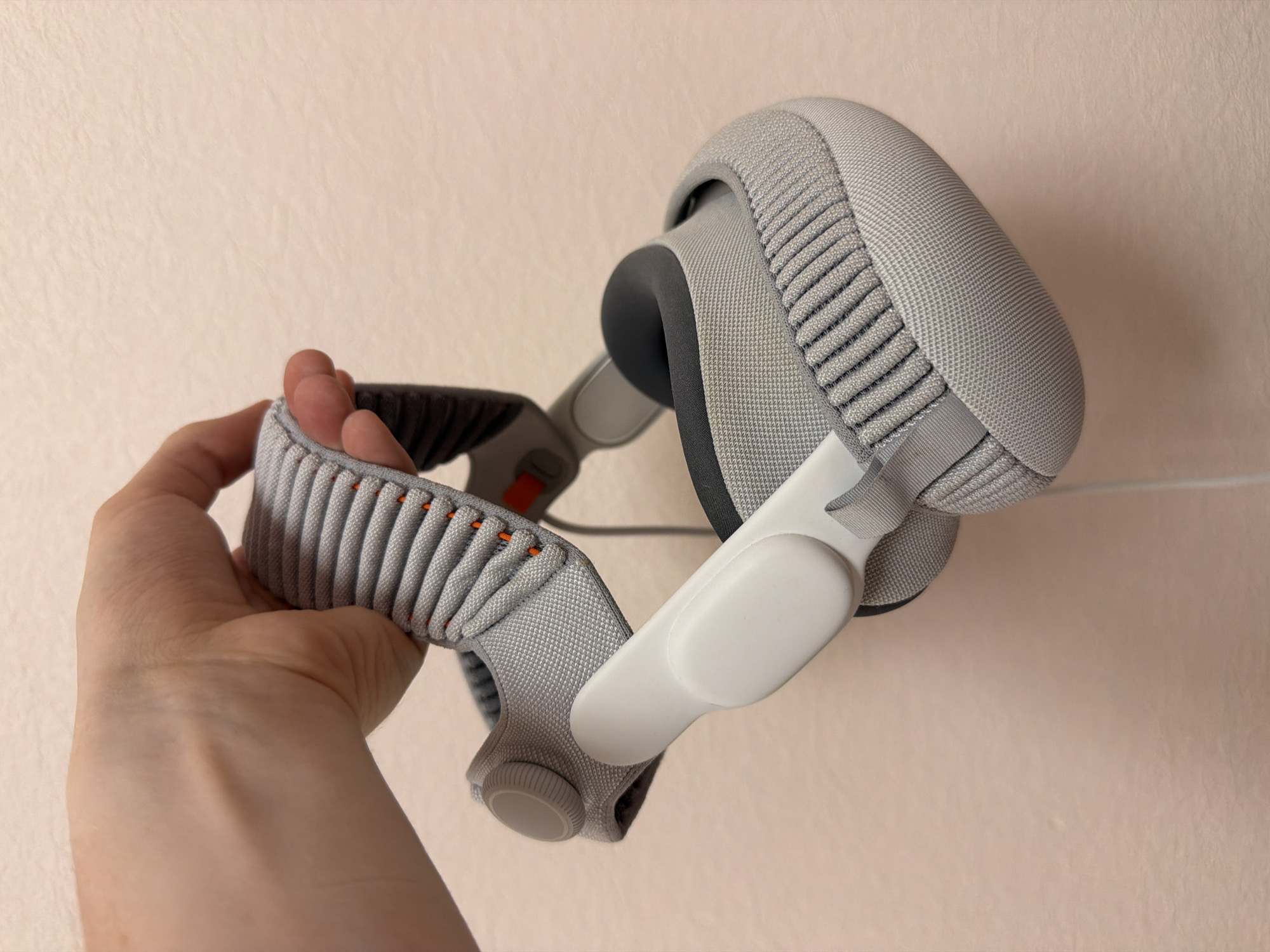 A person's hand is holding a pair of gray headphones with a fabric-covered headband. The headphones have white earcups and a white adjustable arm. The background is a light pink wall.