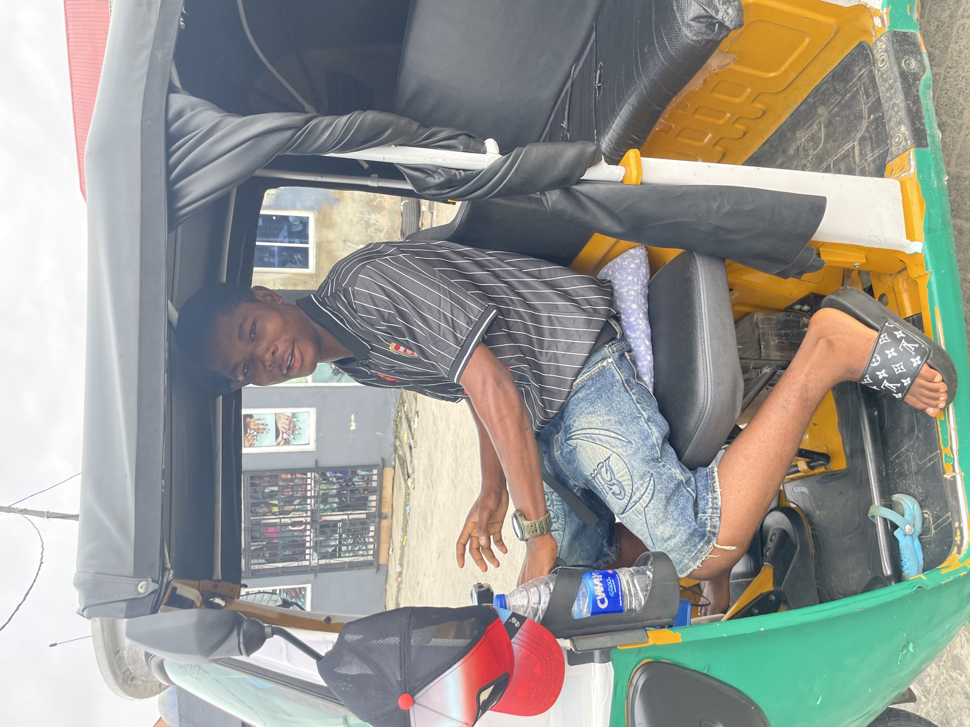 A young Black male, likely a teenager, is seated in the driver's position of a green and yellow auto-rickshaw in Ado, Nigeria. He is wearing a black and white striped polo shirt, ripped denim shorts, and black slide sandals with a Louis Vuitton pattern. On his left arm, he wears a dark-colored smartwatch. A red and black baseball cap is hanging on the side of the driver's seat, and a bottle of "Chivay" water is secured in a holder. The background shows a street scene with buildings, including one with barred windows and posters depicting people's hands. The auto-rickshaw is parked on a dusty, paved surface.