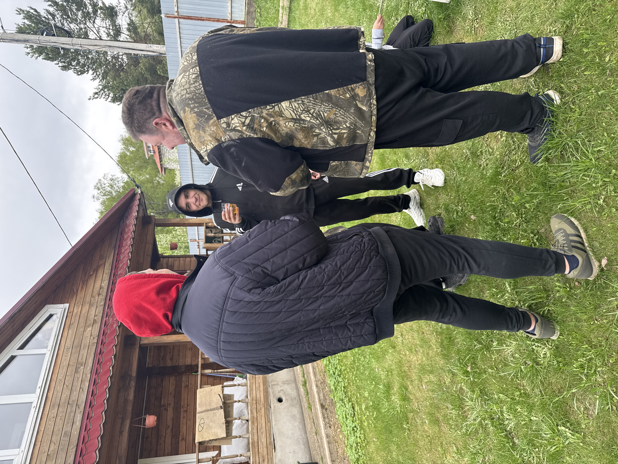 A group of four men are standing in a grassy area in what appears to be a residential or recreational setting, likely in Dmitrov, Russia. The environment includes a wooden house with a red roof and white-framed windows. A man wearing a black jacket with a red hood stands with his back to the camera. In the center, a man in a black Adidas tracksuit holds a glass and smiles. To the right, a man wears a camouflage jacket over a black fleece. A fourth man is partially visible lying on the grass in the background. The weather appears to be overcast. The overall scene suggests a casual gathering or outdoor social interaction.
