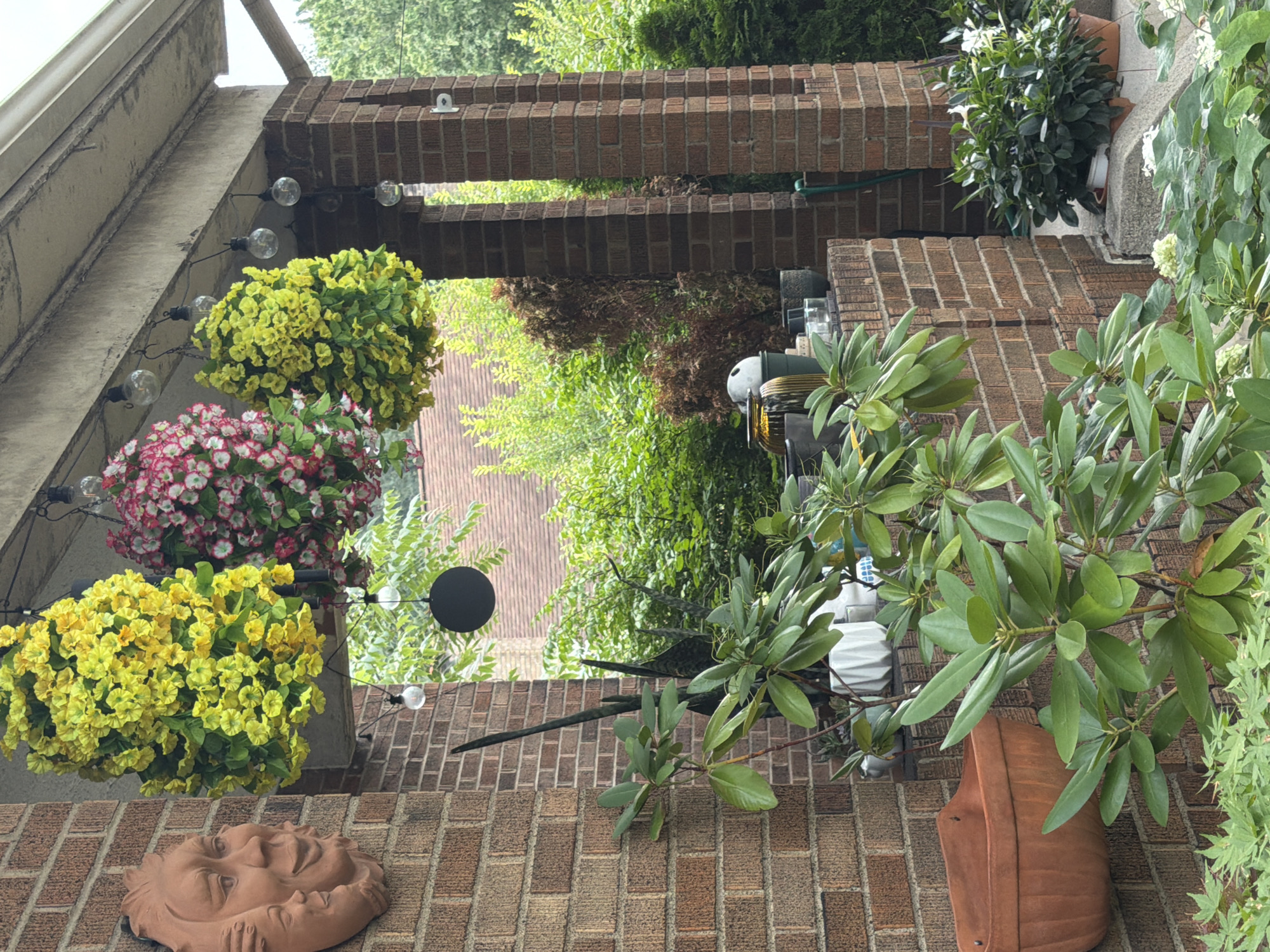 The image captures a detail of a brick house exterior in Pittsburgh, United States of America. In the foreground, lush green foliage, likely from a rhododendron plant, dominates the lower left and center of the frame. To the left, a terracotta wall planter is mounted on the brick wall. Suspended from the overhang above, three large, cascading floral arrangements are visible: two with bright yellow flowers and one with pink and white flowers. Several decorative light bulbs are strung along the overhang, adding to the ambiance. A clay sun face decoration is affixed to the brick wall on the far left. In the background, dense green foliage and trees are visible, suggesting a residential or garden setting. The scene is tranquil and appears to be an outdoor porch or entryway.
