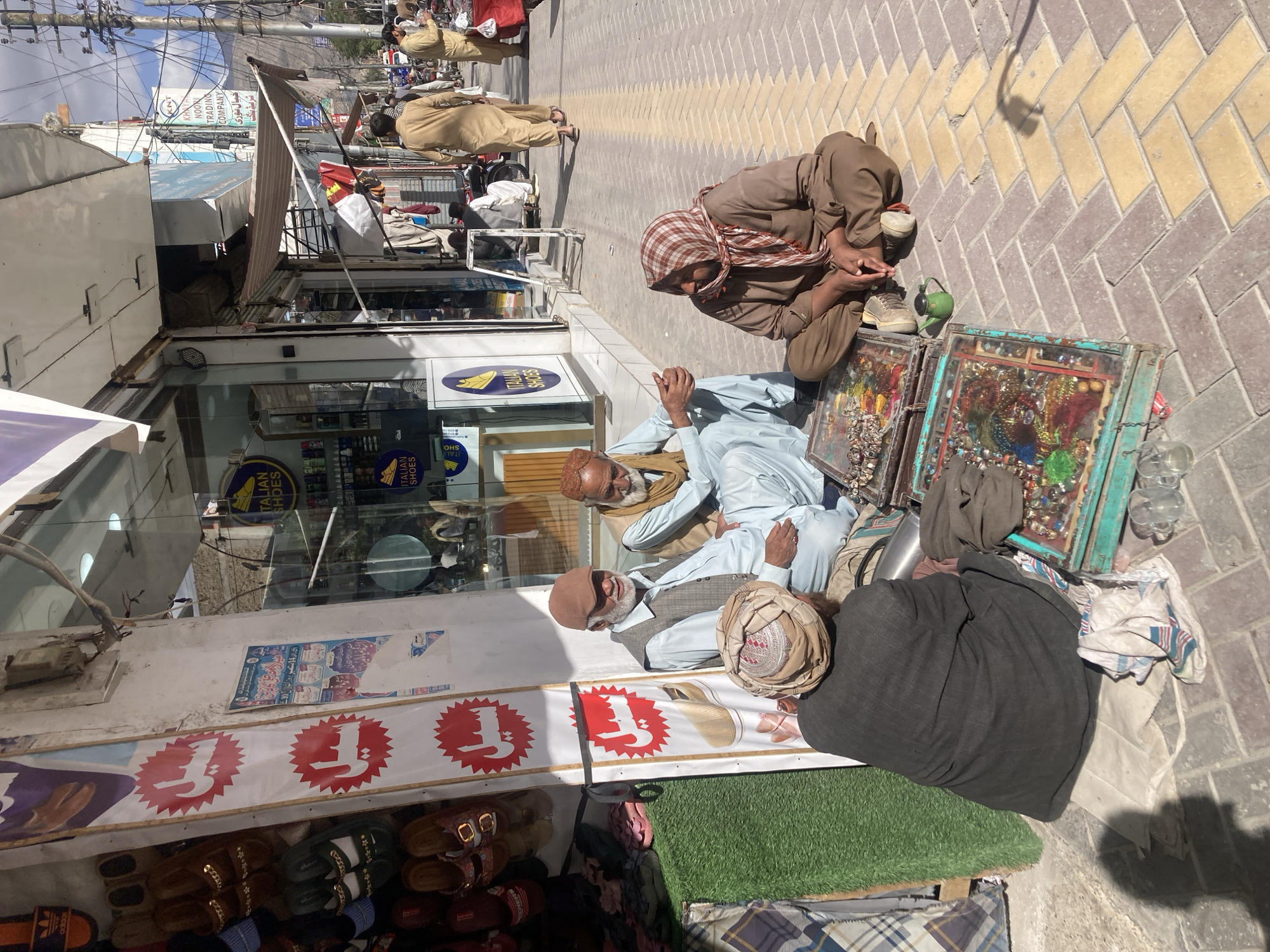 The image shows a street scene in Quetta City Tehsil, Pakistan. On the right side of the image, three men are sitting on the pavement next to what appears to be their wares, two rectangular frames filled with colorful objects. One man is wearing a light blue outfit and a tan hat. Another man wears a checkered headscarf and a brown outfit. The third man is wearing a light blue outfit and a tan hat. Behind them, on the left, is a shop with a shoe display. Several people can be seen walking by, some in traditional Pakistani clothing. The street is paved with alternating tan and gray bricks. Overhead, there are power lines and shop awnings. There is signage with the words "NOOR TRADING COMPANY" and "ITALIAN SHOES."