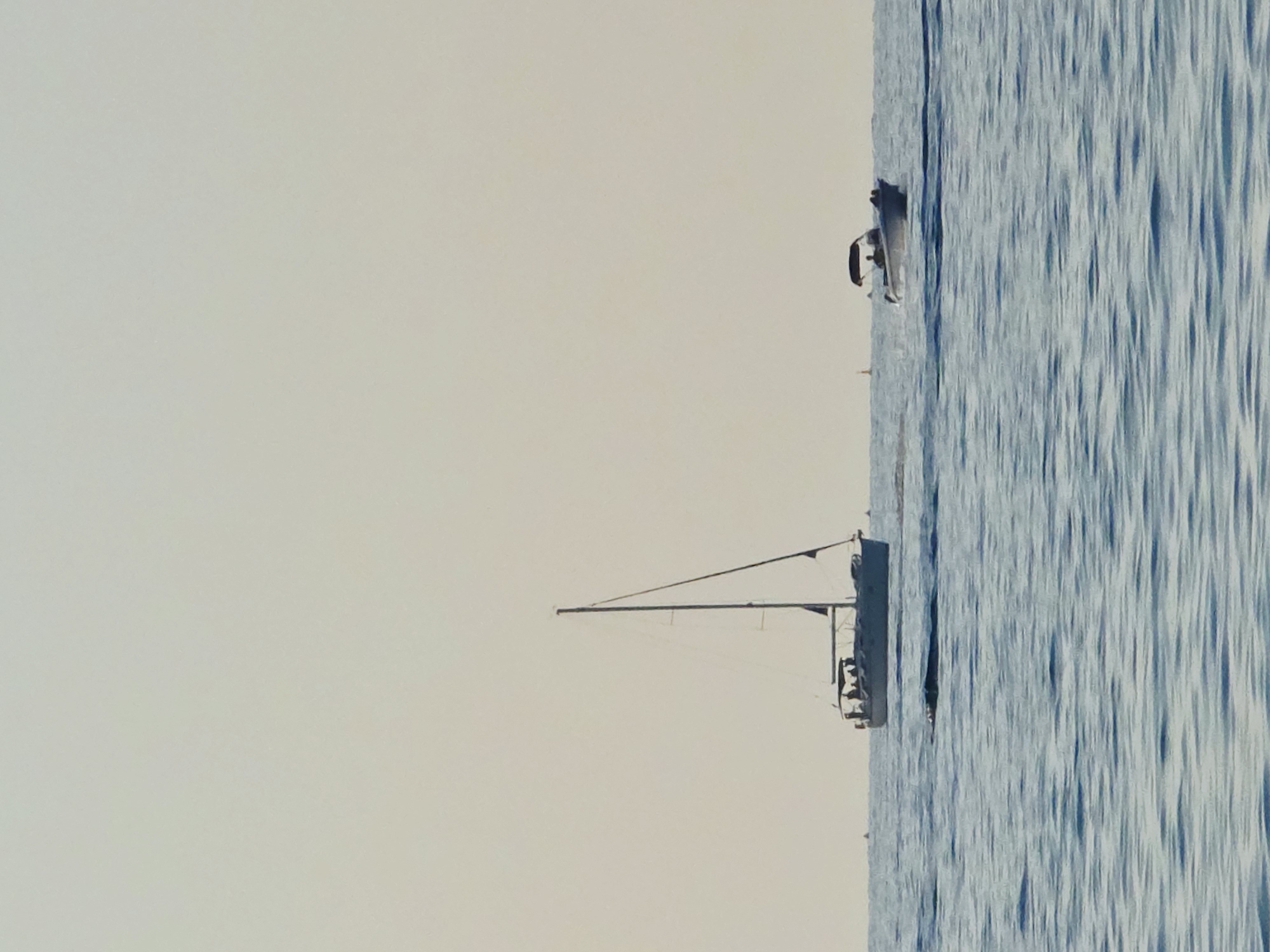 The image depicts a serene seascape with two boats on calm blue waters under a clear, pale sky. On the left, a sailboat with its mast clearly visible is sailing. Several figures can be seen on the deck of the sailboat. To the right, a smaller motorboat with an overhead canopy is moving. People are also visible on the motorboat. In the distance, a small orange buoy can be spotted. The scene is consistent with the coastal waters of Malia Municipal Unit, Greece, characterized by its clear blue sea and typical nautical activities.