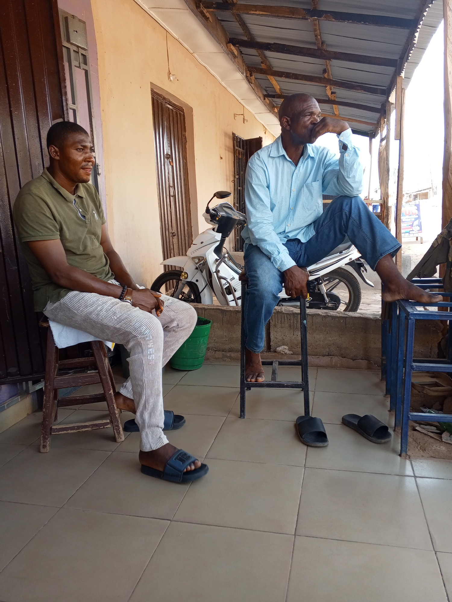 Two African men are seated outside a building in Sabon Tasha, Nigeria. One man, wearing a green polo shirt and striped pants, sits on a wooden stool, looking off to the left. The other man, dressed in a light blue button-down shirt and jeans, sits on a metal stool with one foot resting on a blue metal table, looking thoughtful with his hand on his chin. Behind them, a white motorcycle is parked. Each man has a pair of sandals near their feet. The building has a tan exterior with brown doors and window frames. The area is covered by a simple corrugated metal roof supported by wooden beams.