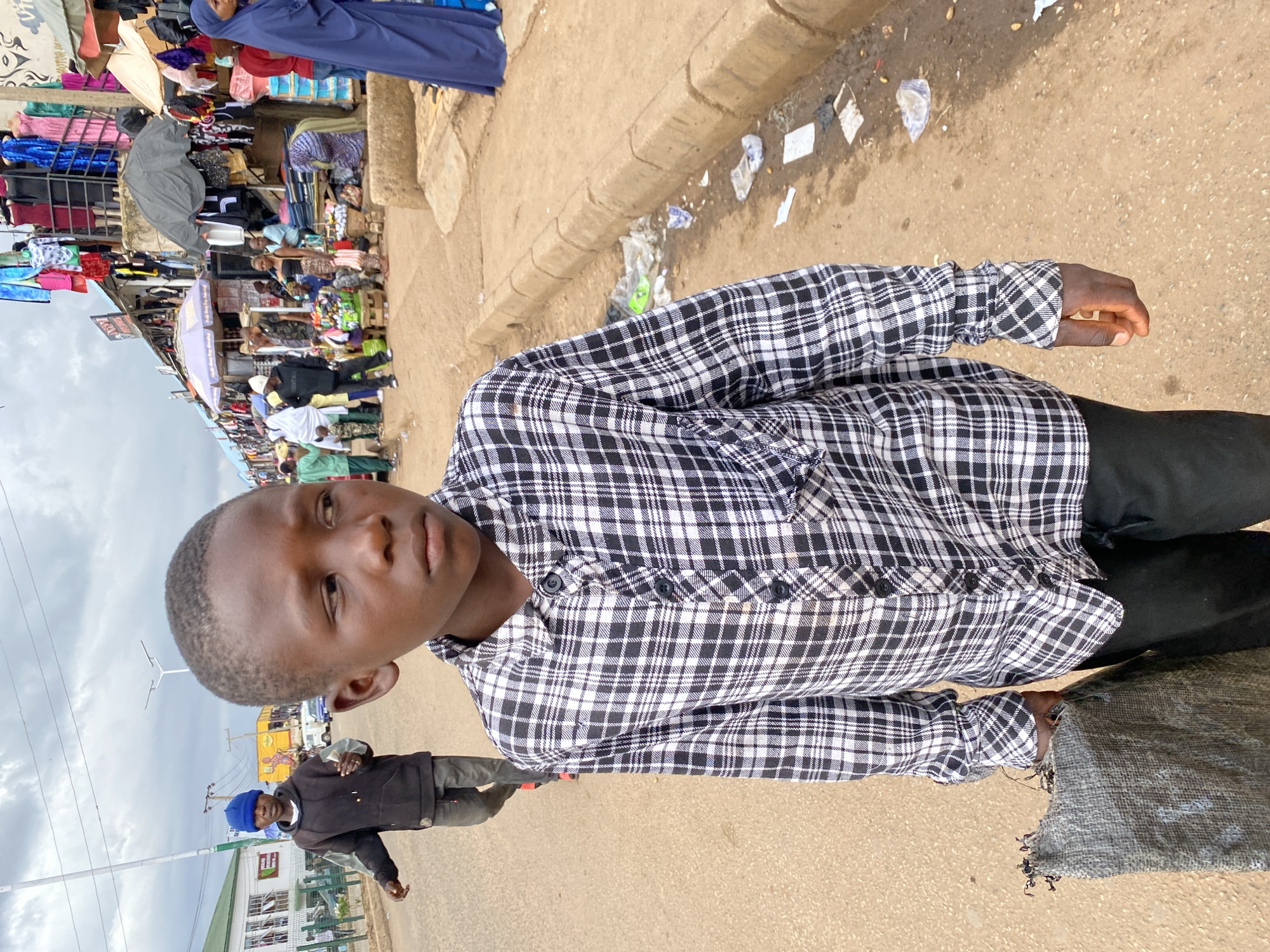 A young boy in a black and white checkered shirt and black pants stands on a dirt road in Bukuru, Nigeria. He is carrying a gray bag in his left hand. In the background, there are market stalls with various goods displayed, including clothing and fabrics. Several people are visible in the market area, some wearing traditional Nigerian attire. The sky is overcast with clouds. The road is lined with shops and appears to be a busy commercial area.