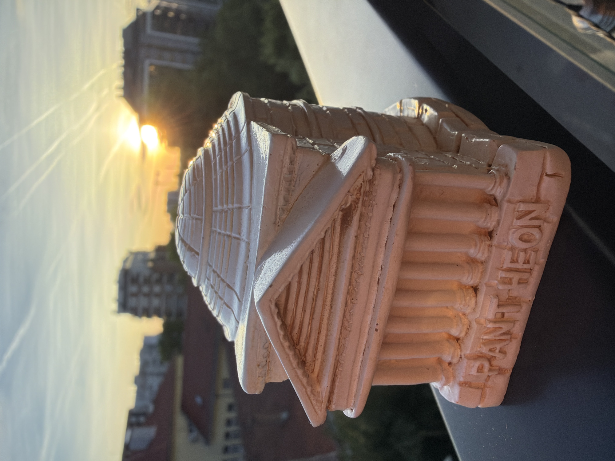 The image shows a miniature Pantheon statue placed on a ledge or windowsill, with a cityscape visible in the background during a sunset or sunrise. The sky is illuminated by the sun, and the silhouette of buildings can be seen. The statue is white with textured details. The word "PANTHEON" is embossed on the statue. The city is of unknown location.