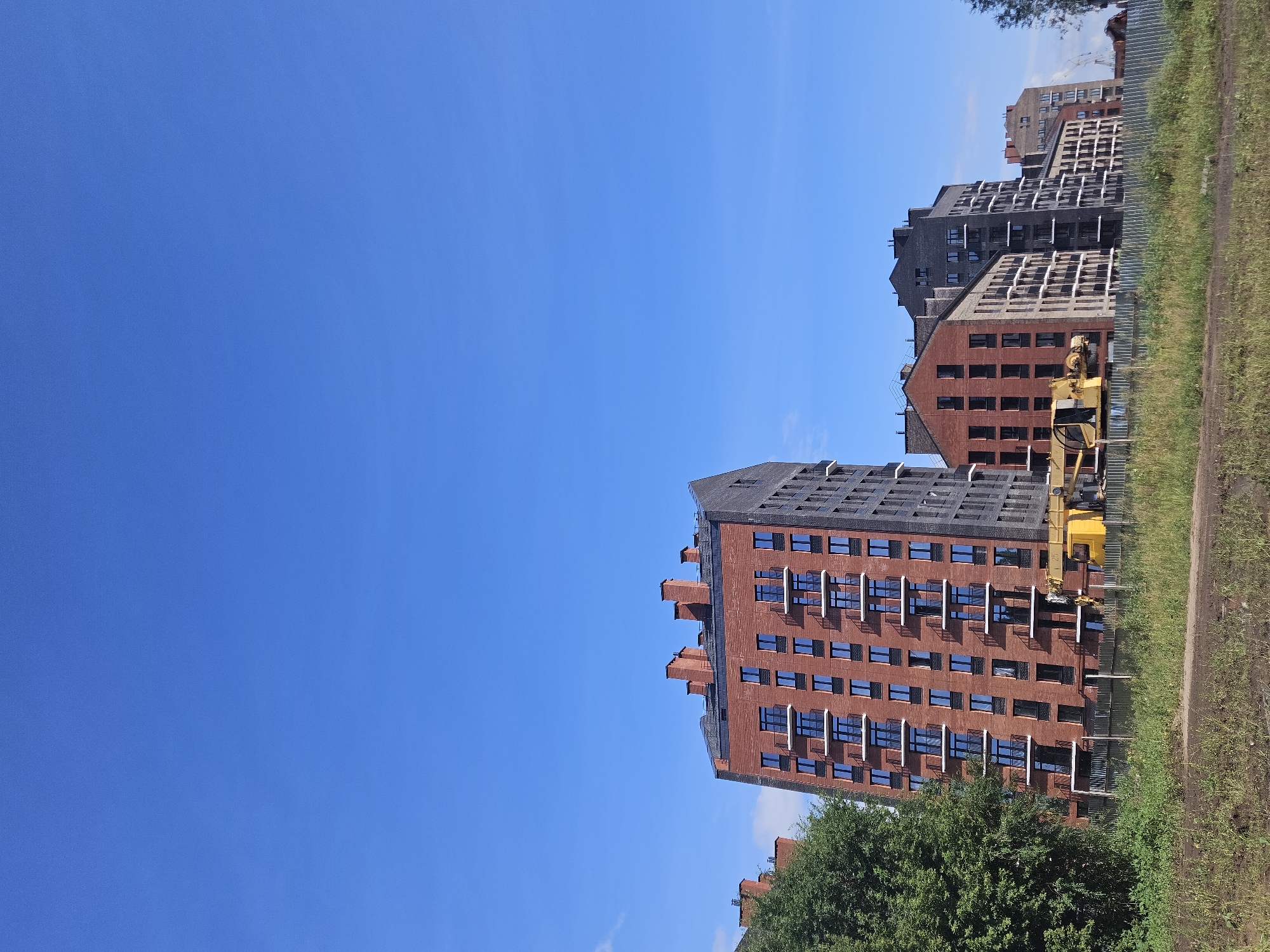 The image depicts a construction site in Ulyanovsk, Russia, featuring several modern apartment buildings under various stages of completion. The buildings are predominantly brick-faced, with some incorporating dark grey cladding. Numerous windows and balconies are visible, indicating residential units. A large yellow construction crane is present in the foreground, positioned behind a metal fence. The sky is a clear, bright blue with a few wisps of clouds. Green trees and overgrown grass are also part of the landscape. The scene suggests ongoing urban development within the city.