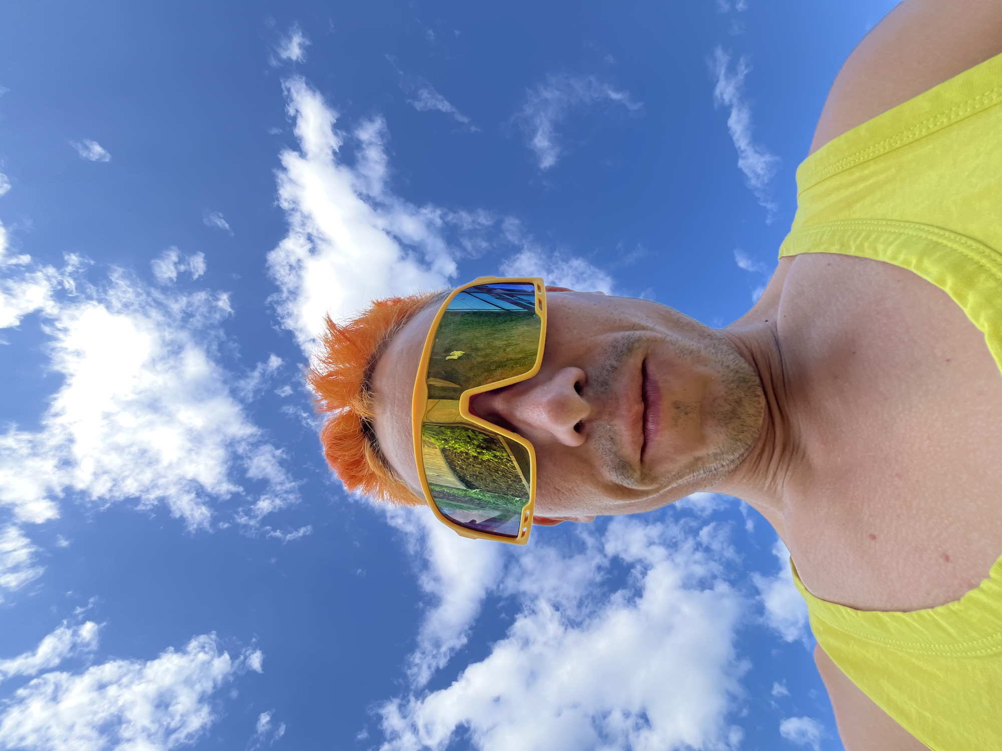 A man with bright orange, spiked hair and yellow sports sunglasses is looking up at the camera. He is wearing a neon yellow tank top. The background is a bright blue sky with scattered white clouds. The image is a low-angle shot, making the sky appear vast. The man's face is visible, with a slight smile. His sunglasses reflect the green foliage and blue sky. The image does not provide any information about the location in the city of Chaikovsky, Russia.