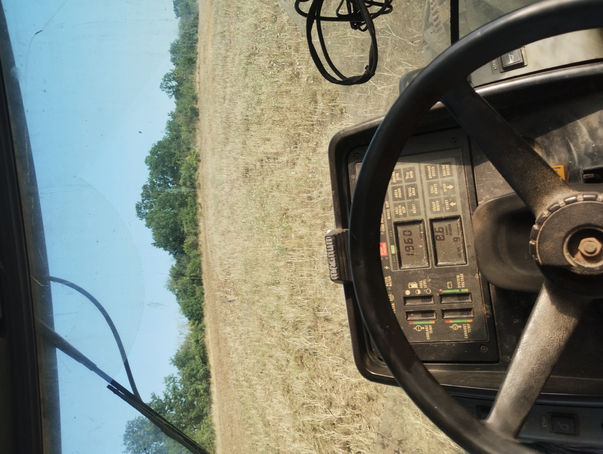 The image is taken from the interior of a tractor cabin, looking out over a harvested field. A steering wheel is prominently featured in the foreground, along with a dashboard displaying various gauges and readouts. The dashboard shows readings for engine RPM, fuel level, coolant temperature, exhaust temperature, and system voltage, along with other agricultural data like area covered and ground speed. Through the windshield, a dry, grassy field extends towards a line of trees in the distance, under a clear blue sky. The scene appears to be rural, likely agricultural land. There is no indication of people or specific activity beyond what is implied by the tractor and field. The location is stated to be Kubadin, Bulgaria, but the image itself provides no direct visual confirmation of this specific locale.