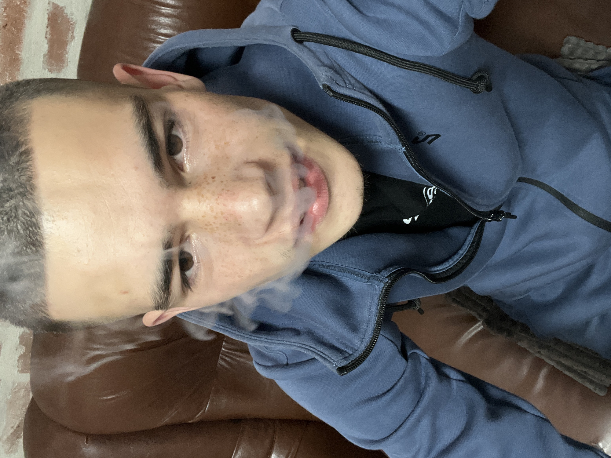 A young man lies on a brown leather couch. He is wearing a blue hooded sweatshirt over a black t-shirt and is emitting smoke from his mouth. The setting appears to be indoors, with a brick wall visible in the background. There is no information to confirm if the location is in Plovdiv, Bulgaria.