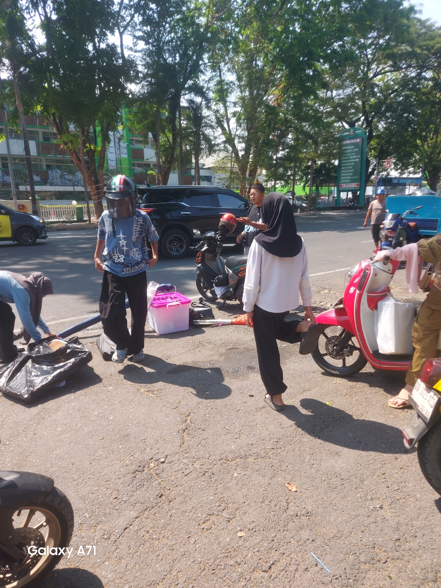 The image depicts a street scene in Makassar, Indonesia, under bright sunlight. In the foreground, a person wearing a helmet with a visor and a blue t-shirt stands facing away from the camera. Next to them, a black plastic bag and a pink container are on the ground. To the left, another person wearing a blue long-sleeved shirt and a hijab is bending down near a black garbage bag. A red and white scooter is parked to the right, with a white bag in its storage compartment. Behind the scooter, a person wearing khaki pants and a patterned shirt is partially visible. In the middle ground, a black SUV is parked, and a motorcycle is also visible. Several trees line the street, and a building with green and white facades is in the background. A large green sign with text is also visible in the background, suggesting an official area or institution. The overall impression is a busy outdoor environment, possibly near a public place or a market.