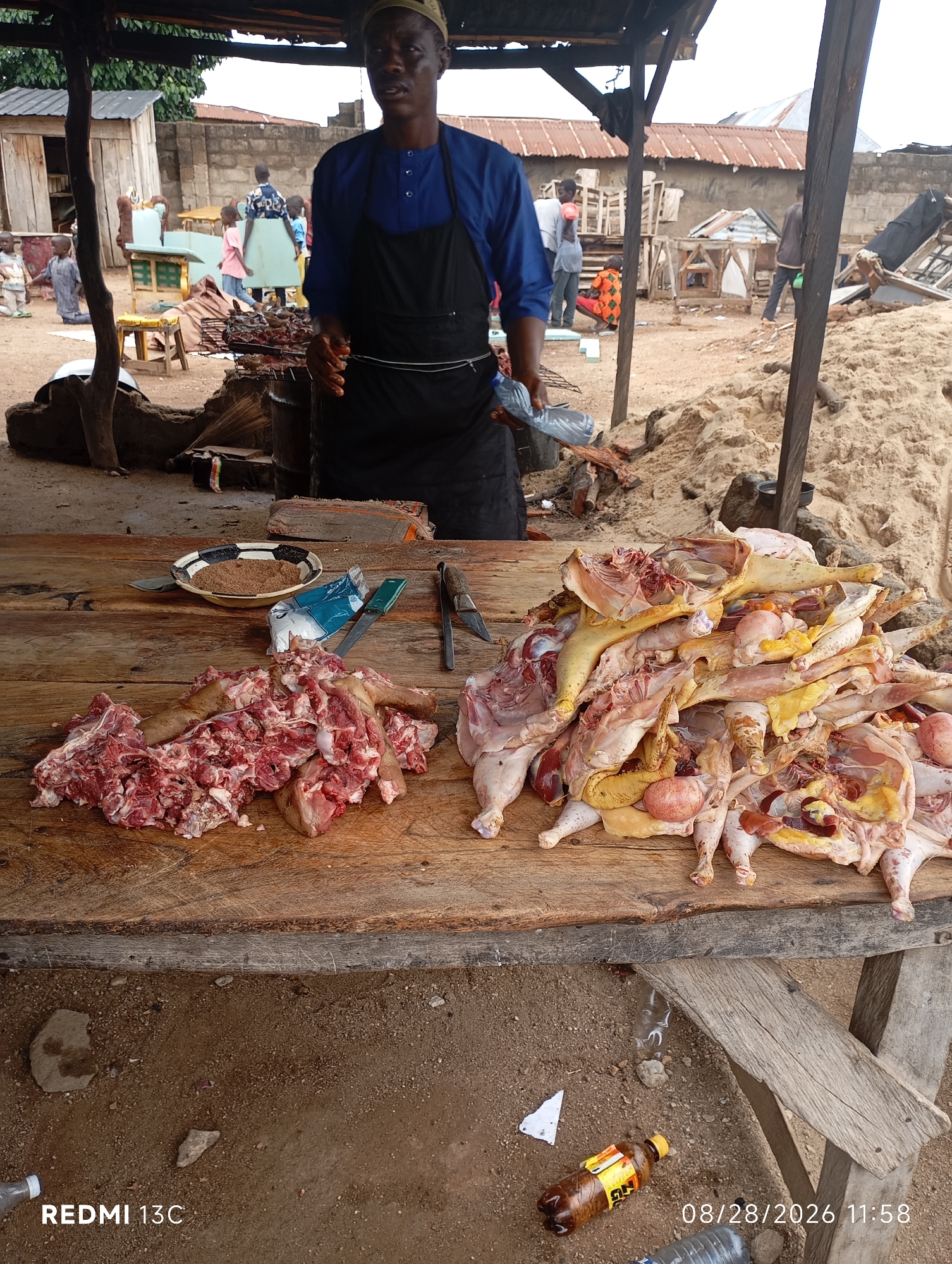 The image depicts an outdoor market scene in Shendam, Nigeria. A man wearing a blue garment and a black apron stands behind a wooden table laden with raw meat. To the left, a pile of red meat, likely pork or beef, is arranged on the table. To the right, a large quantity of cut chicken, including whole birds and parts, is displayed. Also on the table are a bowl of spices, a plastic bag of seasoning, and two knives. In the background, various structures, including wooden stalls and buildings with corrugated iron roofs, are visible. Several other people, including children, are present in the background, some of whom appear to be engaged in market activities. The ground is sandy and dusty, with some debris scattered around. A plastic bottle of beverage is visible on the ground in the lower right corner. The overall scene suggests a lively marketplace where meat is being sold.