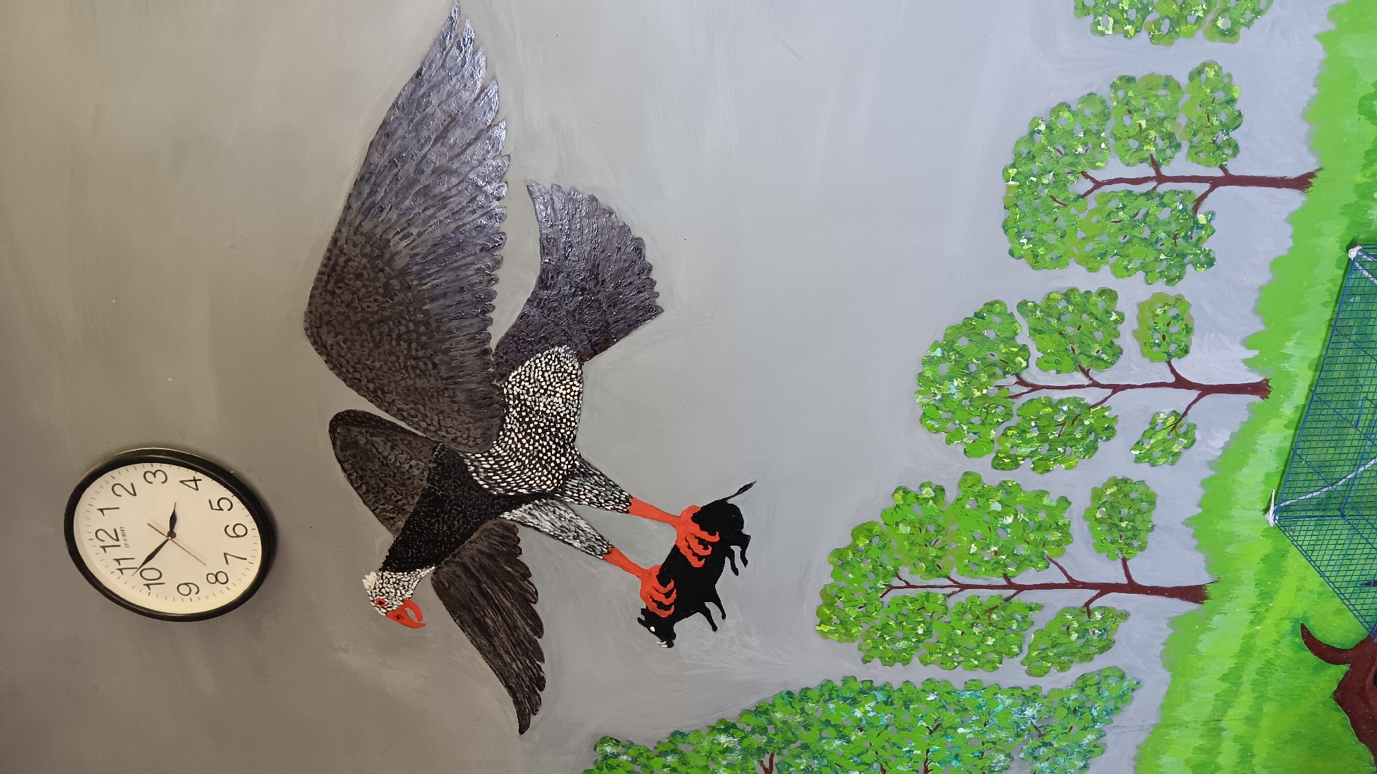 The image is a photograph of a mural painted on a grey wall. The mural depicts a large, stylized bird in flight, holding a small black animal in its talons. The bird has grey and black speckled plumage and bright orange legs and feet. Its head is white with red markings around the eyes and beak. The bird is positioned in the upper half of the image. Below the bird, the mural shows a green, grassy landscape with several stylized trees with round, green canopies. In the lower right corner, a green wire cage is partially visible. In the upper left corner of the image, a white analog clock with black numbers and hands is mounted on the wall. The overall style of the mural is somewhat primitive and colorful. The location is identified as Sonobekel, Indonesia.