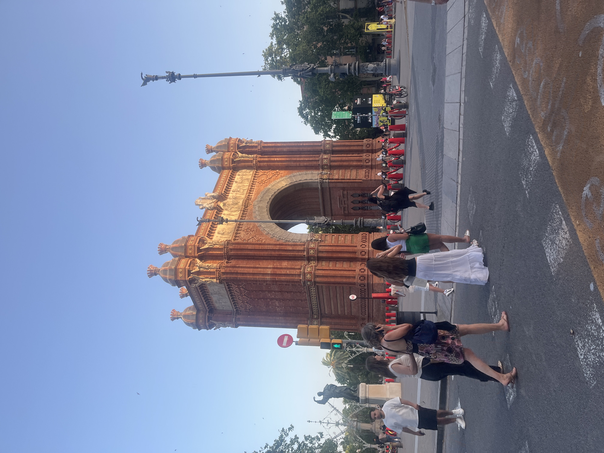 This is a ground-level, medium-shot, eye-level photograph of the Arc de Triomf in Barcelona, Spain. The monument, made of red brick, dominates the background and is bathed in the warm light of a setting sun. The sky is a clear, pale blue, with a few small birds visible.
In the foreground, several people are walking across a street. On the left, a man in a white t-shirt and black shorts walks towards the viewer. To his right, two women walk away from the viewer; one wears a patterned dress and the other a white top and black pants. Further to the right, two more women walk away from the viewer, one in a white maxi skirt and the other in a black top and green shorts.
The street has crosswalk markings and a traffic light system. A statue of a man with outstretched arms stands to the left of the Arc de Triomf, partially obscured by trees and a lamppost. In the middle ground, a line of red-and-white bollards separates the sidewalk from a rental bike station. Trees and buildings are visible in the background, suggesting an urban environment. The overall atmosphere is one of a pleasant late afternoon or early evening in a popular public space.