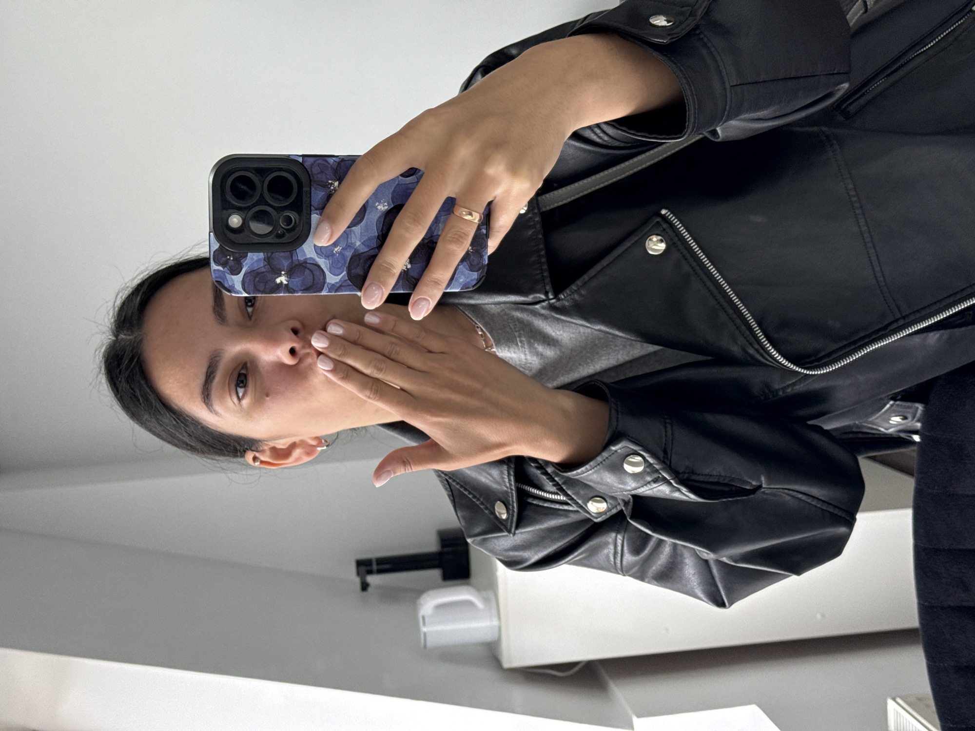 A woman is taking a selfie in what appears to be a bathroom. She is wearing a black leather jacket over a grey shirt. She is holding a phone with a blue floral case. She is puckering her lips and holding her hand up to her face as if blowing a kiss. Her nails are painted a light, neutral color and she is wearing a plain gold ring on her left ring finger.
The background is a standard bathroom with white walls and fixtures. A towel rack with a white paper towel dispenser is visible. The location in the city of Chaikovsky, Russia cannot be determined from the image.