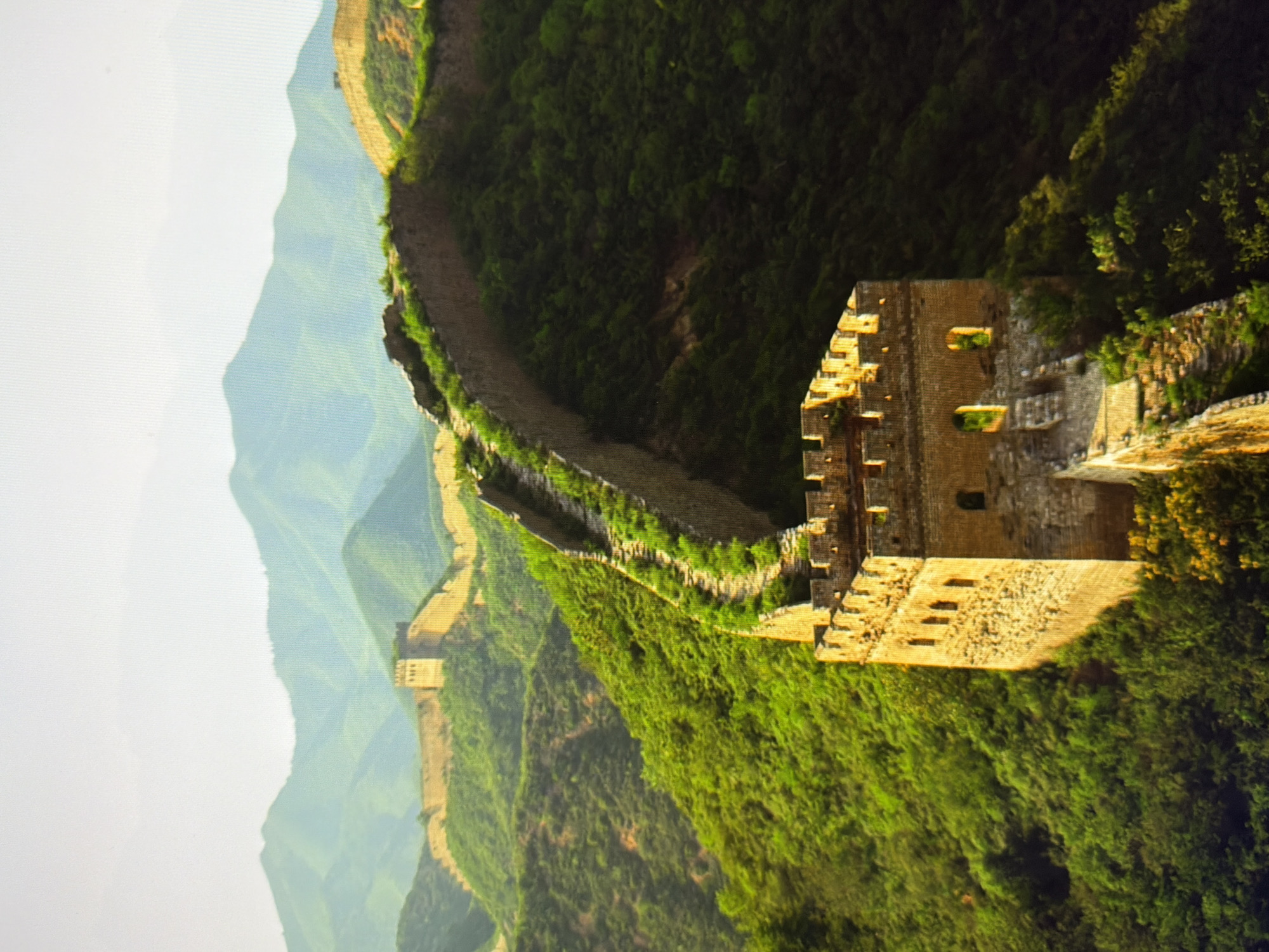 The image displays a section of the Great Wall of China snaking along lush, green, mountainous terrain under a bright, hazy sky. In the foreground, a stone watchtower with crenellations and arched openings stands prominently. The wall itself, constructed from rough stone blocks, follows the contours of the steep hills, disappearing into the distance where more sections of the wall and other watchtowers are visible against a backdrop of layered, blue-green mountains. The scene is devoid of people or any urban elements, focusing solely on the ancient structure and its natural surroundings. There is no indication that this scene is located in Kyiv, Ukraine.