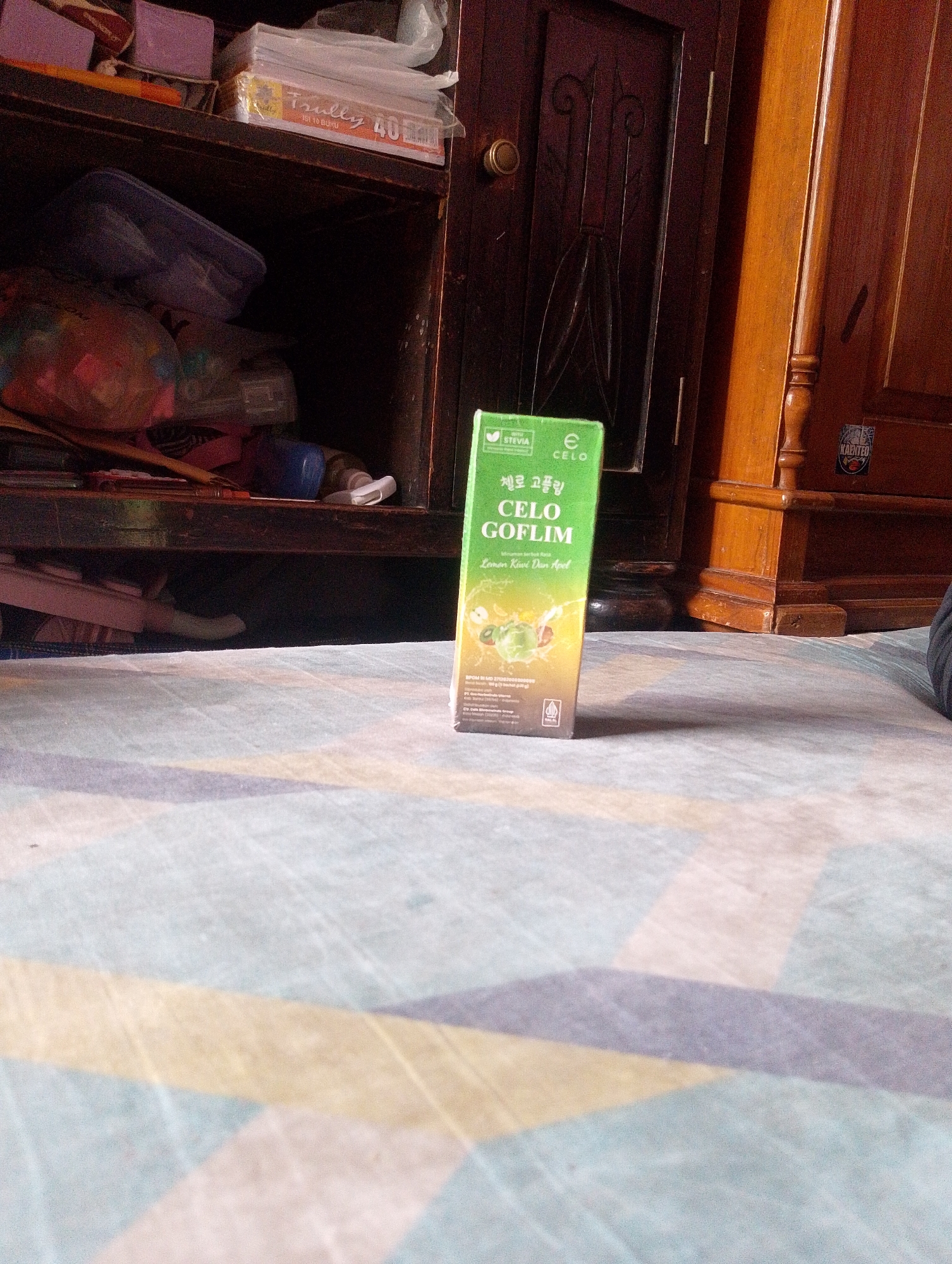 The image shows the interior of a room, likely a home, with wooden furniture and shelves. On the top shelf, there are several items including what appears to be a stack of notebooks with "Tulloy 40" printed on them. Below, on a lower shelf, are various items in plastic bags, and on the floor in front of the furniture, a green box of "CELO GOFLIM" with "Lemon Kiwi Dan Apel" flavor is prominently displayed. The floor is covered with a patterned rug. The lighting suggests an indoor setting, possibly daytime given the light sources. There are no people or animals visible in the image. The overall impression is of a personal space, possibly a storage area within a residence.