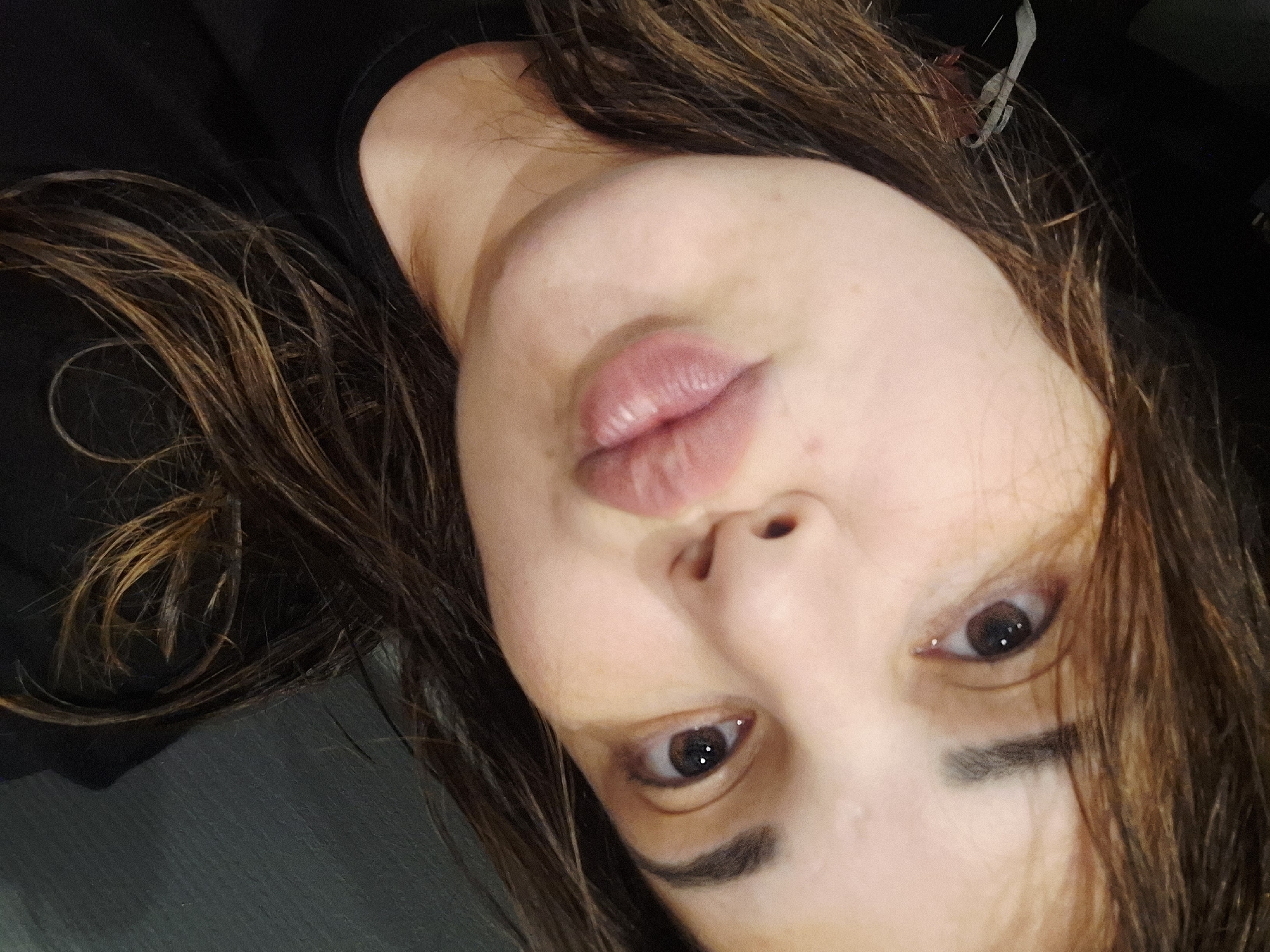 A close-up selfie shows a woman with her head tilted, looking directly at the camera. Her hair, a mix of brown and lighter highlights, falls across her shoulders and frames her face. Her lips are slightly puckered in a "duck face" expression. Her dark brown eyes are open, with light reflecting in the pupils. One visible eyebrow is dark and well-defined. Her skin appears fair, with a few subtle blemishes visible. The background is mostly dark and indistinct, suggesting an indoor setting. There is no visible text. The time of day cannot be determined from the image.