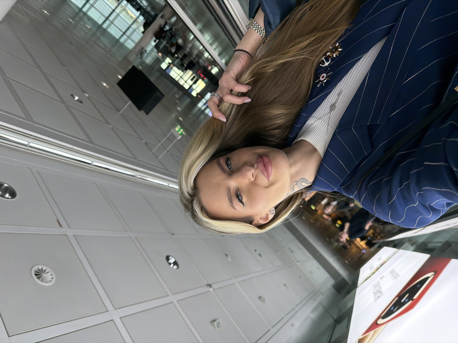 A fair-skinned female subject is depicted in a selfie taken within Munich Airport (MUC) in Freising, Germany. She has long, straight blonde hair extending over her left shoulder, with her left hand lightly touching it. Her face shows defined eyebrows, dark eyeliner, and full, slightly pursed lips. A tattoo resembling a stylized 'S' or snake is visible on the right side of her neck. She is dressed in a navy blue pinstriped blazer worn over a white ribbed top. Accessories include "CD" logo stud earrings, a silver-toned necklace, and two brooches on her blazer's lapel (one appearing as a ship's wheel with a red center, the other as an anchor). Her left hand bears multiple rings on the ring finger, and her left wrist displays a silver-toned metal link watch and a black elastic hair tie. The background reveals a modern airport terminal interior, featuring a white paneled ceiling with recessed lighting and air vents, large glass windows providing a blurred view of an exterior or distant terminal section, a visible escalator or moving walkway on the right, and a suspended black rectangular digital display. A partially visible white advertising stand for "HUAWEI MATE" is present on the lower left. Blurry figures of other individuals are discernible in the deeper background.