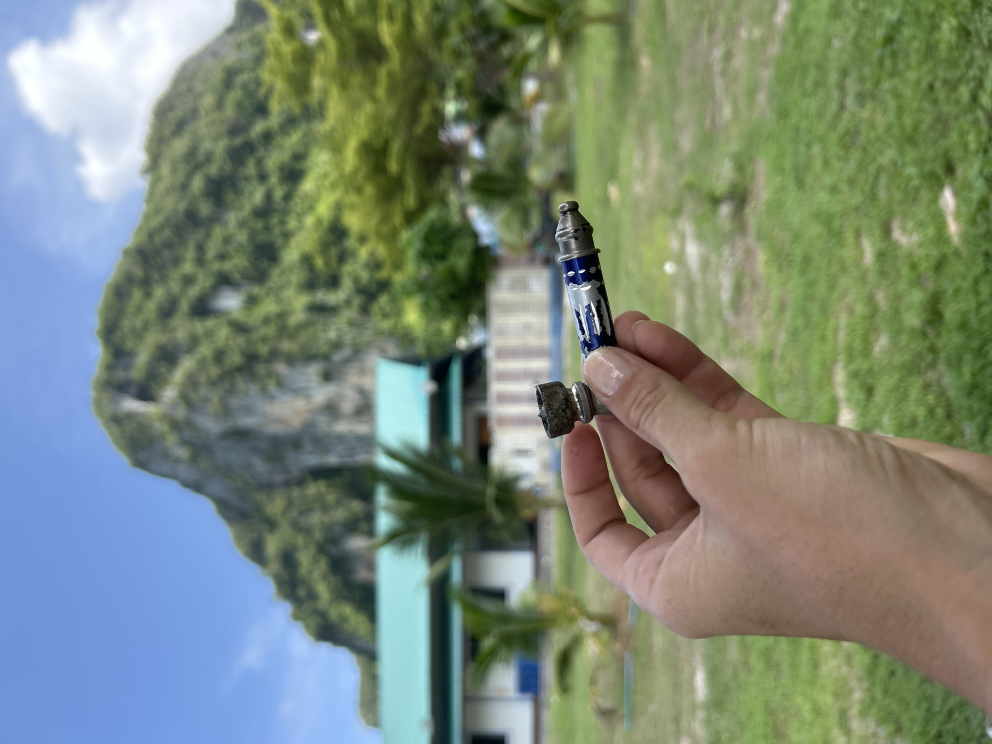 A person's hand holds two parts of a disassembled metal smoking pipe outdoors in Ao Nang, Thailand. The pipe is silver and blue and appears to be used. In the background, a building with a turquoise roof and a large, green, mountainous island under a blue sky with scattered clouds are visible. The foreground consists of a grassy lawn with a few palm trees.