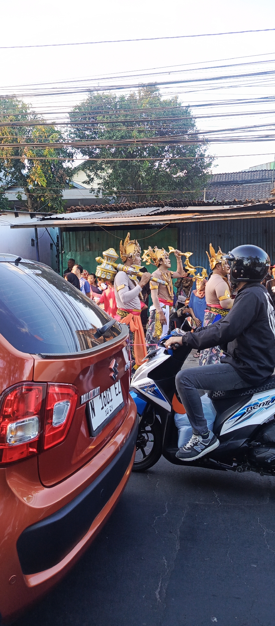 In Malang, Indonesia, a vibrant street scene unfolds as a parade or cultural event takes place. The image captures a procession of individuals in traditional attire, adorned with elaborate golden headdresses and carrying symbolic props. The figures are dressed in richly patterned fabrics, with some wearing orange and pink sarongs, while others sport brown tops and intricate waistbands. Their costumes suggest a ceremonial or performance context, possibly depicting mythological characters or historical figures.
In the foreground, a red Suzuki car, identified by its license plate "N 1601 EZ," is parked, its rear visible. To the right, a person on a blue and white Honda Beat scooter, wearing a black helmet and dark clothing, observes the event. The scooter's license plate is partially obscured. The background reveals a row of modest buildings with corrugated metal roofs, suggesting a commercial or residential area. Overhead, a dense network of electrical wires crisscrosses the sky, a common sight in urban Indonesian landscapes. Trees with green foliage line the street, adding a touch of nature to the urban setting. The overall atmosphere appears lively and festive, with onlookers present in the background, hinting at community participation in the event. The lighting indicates it is daytime, likely late afternoon given the warm, soft light.