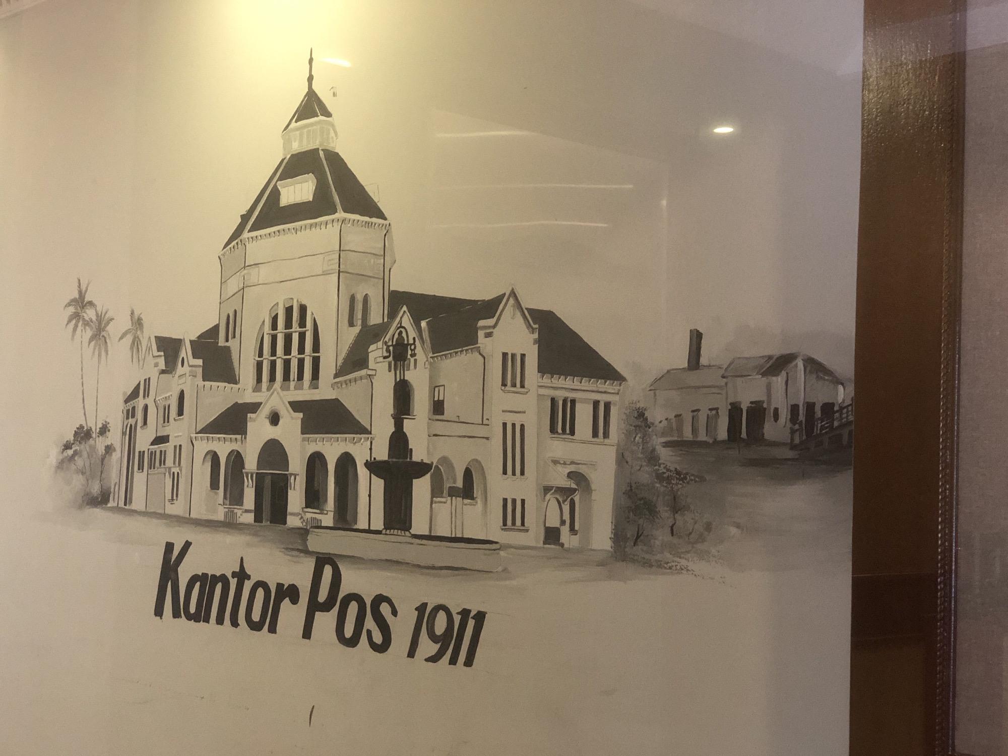 The image is a black and white drawing of the "Kantor Pos 1911" in Medan, Indonesia. The drawing depicts a large building with a central tower, palm trees to the left, a fountain in front, and smaller buildings in the background. The text "Kantor Pos 1911" is written in the foreground. The drawing appears to be displayed in a frame.