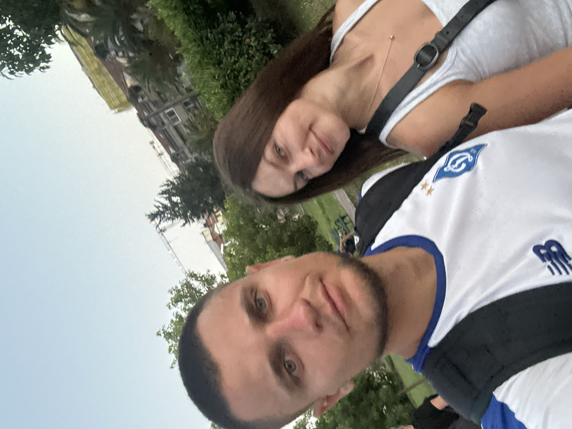 A close-up selfie shows a man and a woman outdoors in the city of Budva, Montenegro. The man, in the foreground, is wearing a white t-shirt with blue trim and a black backpack. He has short dark hair and light eyes. The woman, to his right and slightly behind him, has long brown hair and is wearing a white tank top with a black strap across her shoulder. She is smiling. In the background, there are green bushes, trees, buildings, and a construction crane. The sky is clear and light blue.