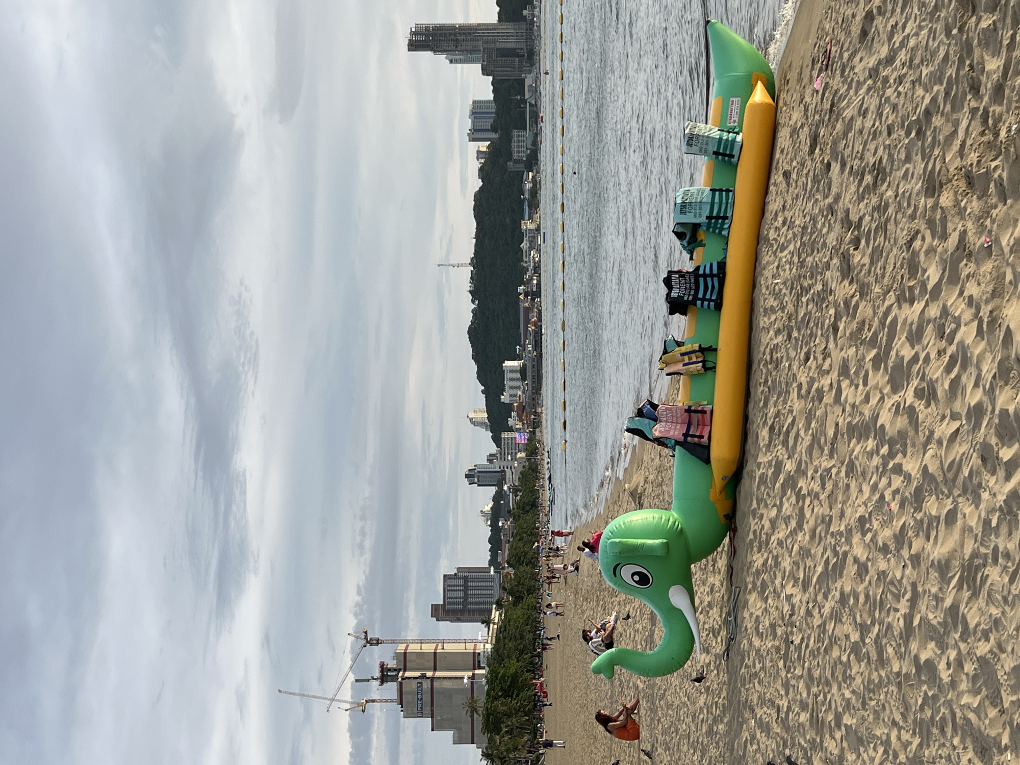 The image depicts a beach scene in Pattaya City, Thailand, on a cloudy day. In the foreground, an inflatable banana boat shaped like a green elephant is resting on the sand near the waterline. Several life jackets are placed on top of the banana boat. The beach is sandy with some scattered debris. In the middle ground, the ocean is visible with a yellow floating barrier marking a swimming area. Further in the distance, the city skyline of Pattaya is apparent, with numerous buildings and construction cranes. People are seen scattered across the beach, some sitting and others walking. The overall atmosphere is that of a typical tourist beach with recreational activities.