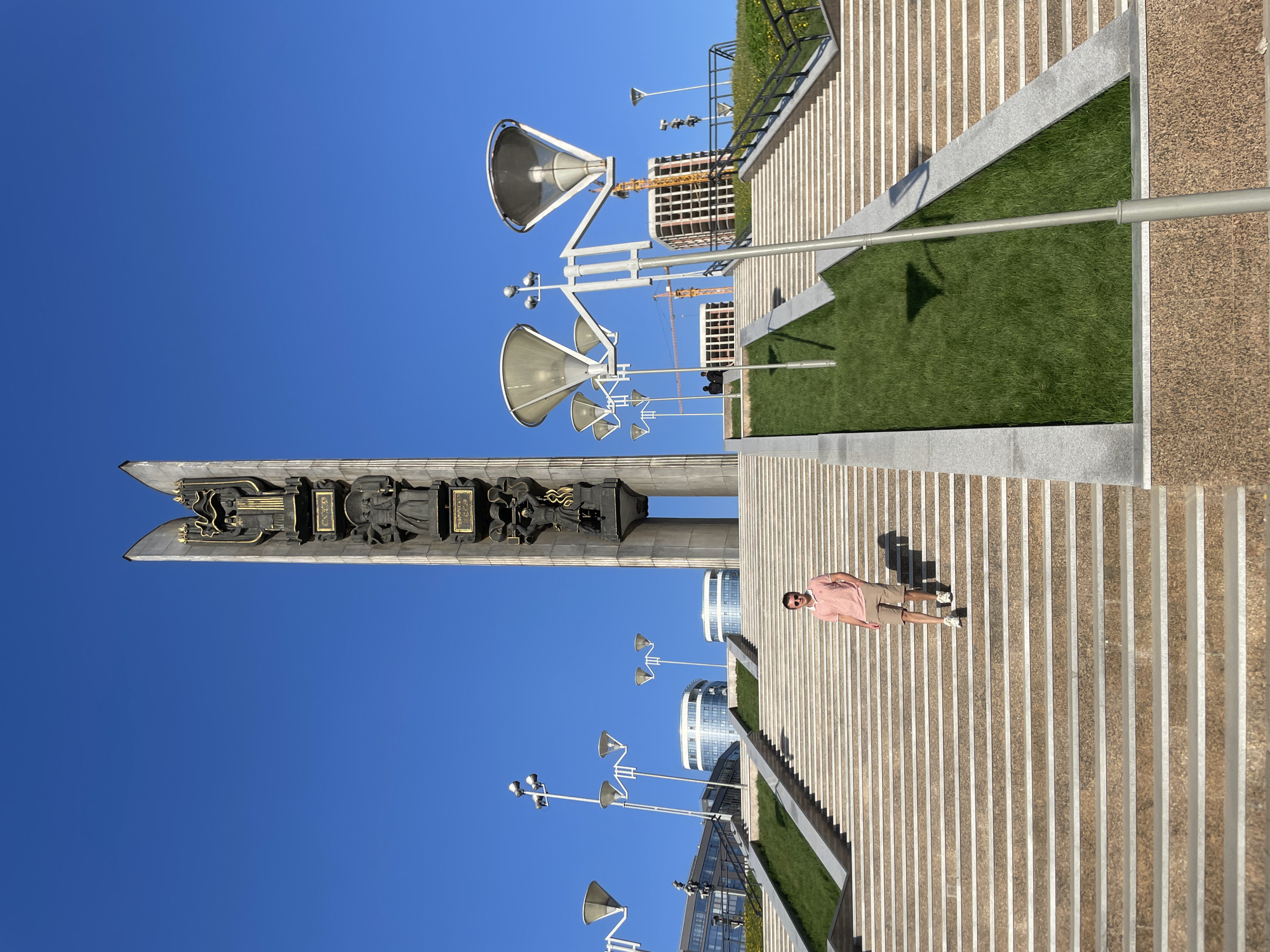 The image shows a man standing on a set of wide, light-colored stone steps in Izhevsk, Russia. In the background, a large obelisk with bronze sculptures and the bright blue sky is visible. Several tall light poles with large, cone-shaped shades are also present. The man is wearing a pink shirt and tan shorts. A patch of green grass is located next to the steps. In the distant background, some buildings are under construction, with cranes visible.