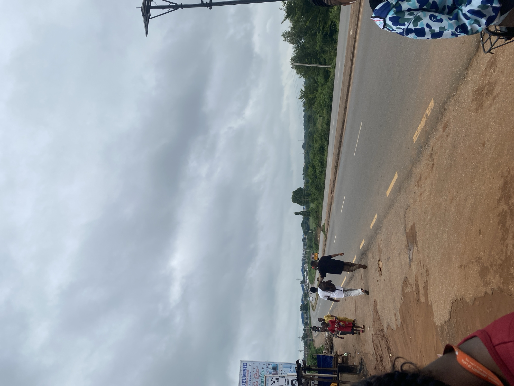 The image shows a street scene in Lafia, Nigeria, under a cloudy sky. A paved road runs horizontally across the image, with a dirt shoulder to the left. Several people are walking along the shoulder, including a group of children and two men. One man is wearing a white shirt and the other is wearing a black shirt and carrying a backpack. Further down the road, a yellow vehicle is visible. On the left side of the image, a billboard advertising "UZUMISHI EVENTS" is present, along with a small roadside stall. Trees and vegetation line the opposite side of the road. A lamppost is visible on the far right of the image.