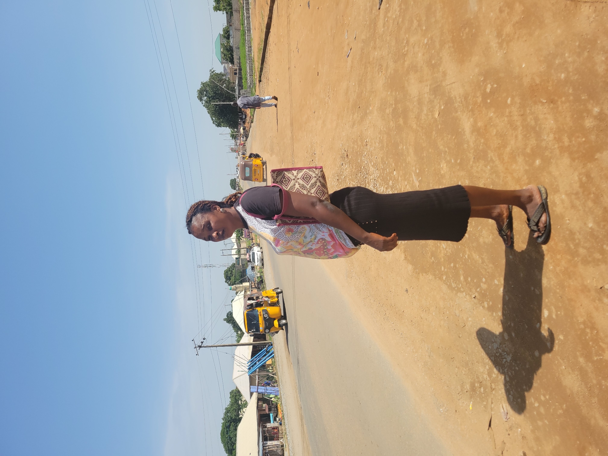 In Kwamba, Nigeria, a woman stands on the side of a dusty road, her shadow cast to her left. She wears a patterned t-shirt, a black skirt, and sandals. A woven bag hangs from her shoulder. The road is unpaved on the side where she stands, transitioning to a paved surface with traffic. A yellow auto-rickshaw is seen driving on the road, and another vehicle, possibly a car, is parked further down. Several buildings with corrugated metal roofs line the road in the background, along with trees and utility poles. The sky above is clear and blue.
