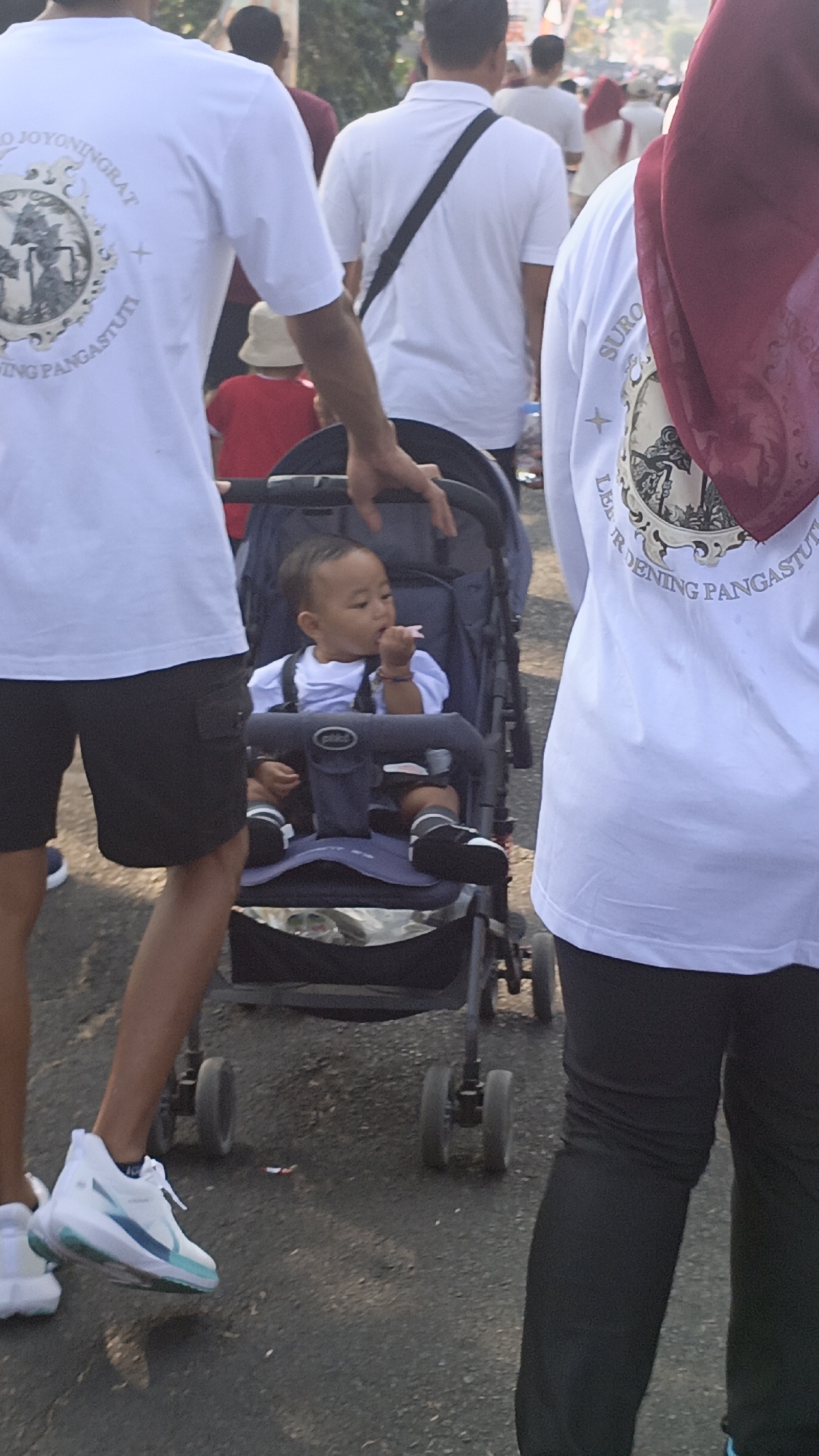 The image captures a daytime street scene in Salatiga, Indonesia, focusing on a baby in a stroller and several individuals walking. In the foreground, a baby with dark hair is seated in a dark blue stroller, facing forward and holding something pink to its mouth. The baby wears a light blue shirt and dark shorts, with black shoes and socks. To the left, a person wearing a white t-shirt with a circular emblem and the text "JOYONINGRAT" and "PANGASTUTI" is seen from the back, their right arm resting on the stroller's handle. They are wearing black shorts and white athletic shoes with blue and green accents. To the right, another person is visible from the back, wearing a white t-shirt with a similar circular emblem and the text "SUBUR" and "RUDENING PANGASTUTI," along with a maroon headscarf. This individual is wearing black pants. In the background, other people are walking, some in white shirts and others in darker clothing. A child wearing a red shirt and a light-colored hat is also visible in the middle ground. The ground appears to be asphalt or a similar paved surface, with some debris scattered. The lighting suggests it is daytime with ambient sunlight.