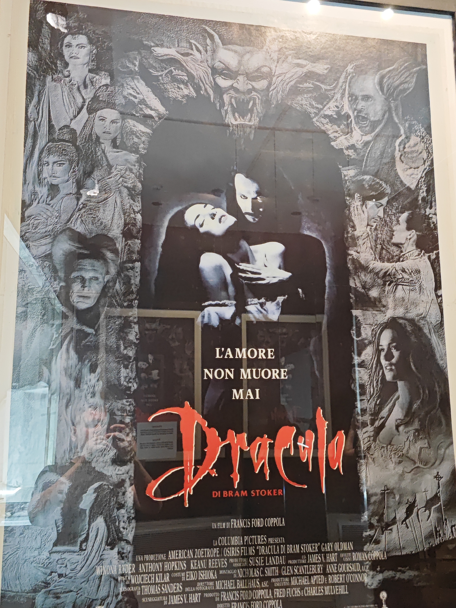 The image is a vertical, slightly angled, eye-level, full shot of a movie poster displayed on a wall in Turin, Italy. The poster is for the 1992 film "Bram Stoker's Dracula." The poster features a dark, gothic design with a large, stylized red title "Dracula" prominently displayed. Around the title, there are illustrations of various characters, including a distressed couple in the center, a monstrous face at the top, and multiple figures, some appearing to be women with elaborate hairstyles, along the sides. The poster also includes text detailing the film's credits, such as the director Francis Ford Coppola, and actors like Winona Ryder, Gary Oldman, and Anthony Hopkins. The poster itself appears to be in good condition, though there are some minor scuffs and reflections on its surface, possibly from the glass protecting it or ambient lighting. The background suggests an indoor environment, likely a cinema or a shop exhibiting movie memorabilia, given the context of a poster display. The reflections visible on the poster suggest the presence of lights and potentially other objects in the vicinity, but no people are clearly depicted in the reflection.