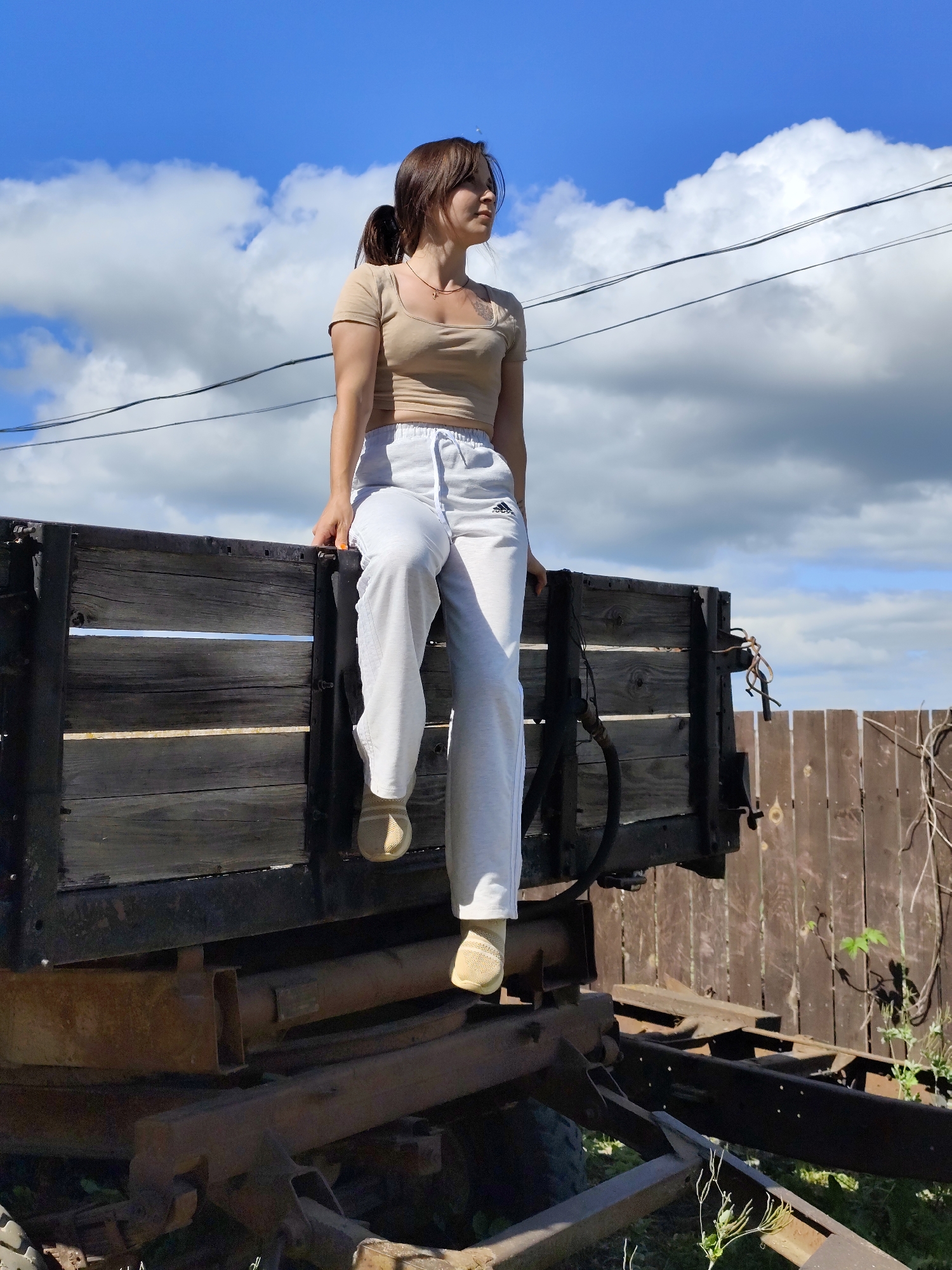 In the city of Fokino, Russia, a young woman with brown hair pulled back in a ponytail is sitting on the edge of a wooden trailer under a partly cloudy blue sky. She is wearing a tan crop top and white sweatpants with an Adidas logo. She is looking off to the right, with her arms resting on the trailer. The trailer is made of weathered wood and metal. In the background, there is a wooden fence and some dry vegetation. The overall impression is one of a rural or industrial setting on a bright, sunny day.