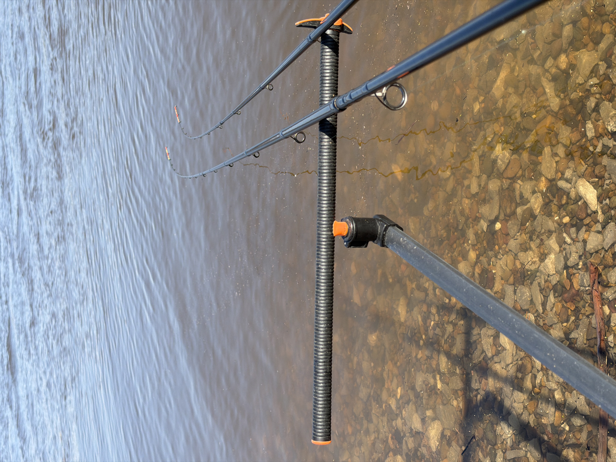 The image depicts two fishing rods placed on a black and orange rod rest, positioned at the edge of a body of water. The left side of the frame shows water with slight ripples, while the right side reveals a rocky shoreline with small pebbles. The location is identified as the city of Chaikovsky, Russia.