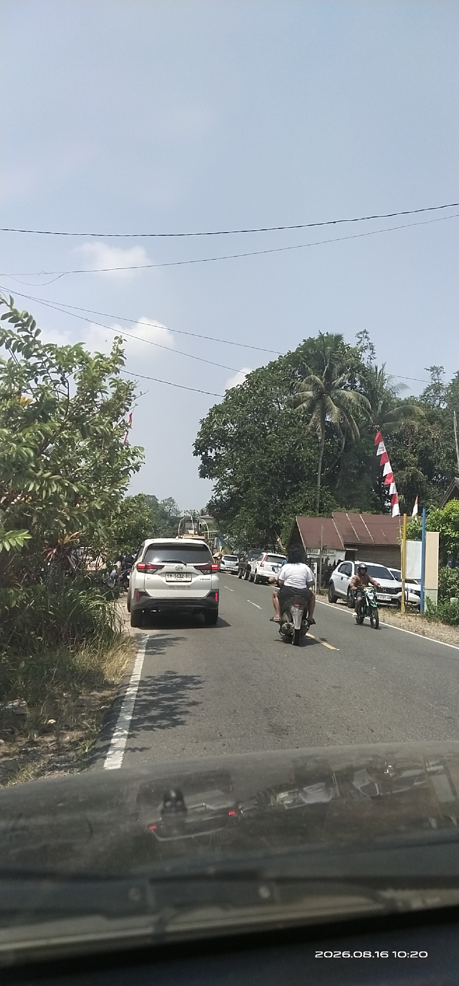 A white SUV is driving on a road in a rural area of Lenggang, Indonesia. The SUV has a license plate that reads "KH 1432 DG". There are trees and vegetation on either side of the road. There are also a few other vehicles on the road, including a motorcycle with two people on it and several other cars. The sky is blue with some clouds. The image appears to have been taken on August 16, 2026, at 10:20 AM.